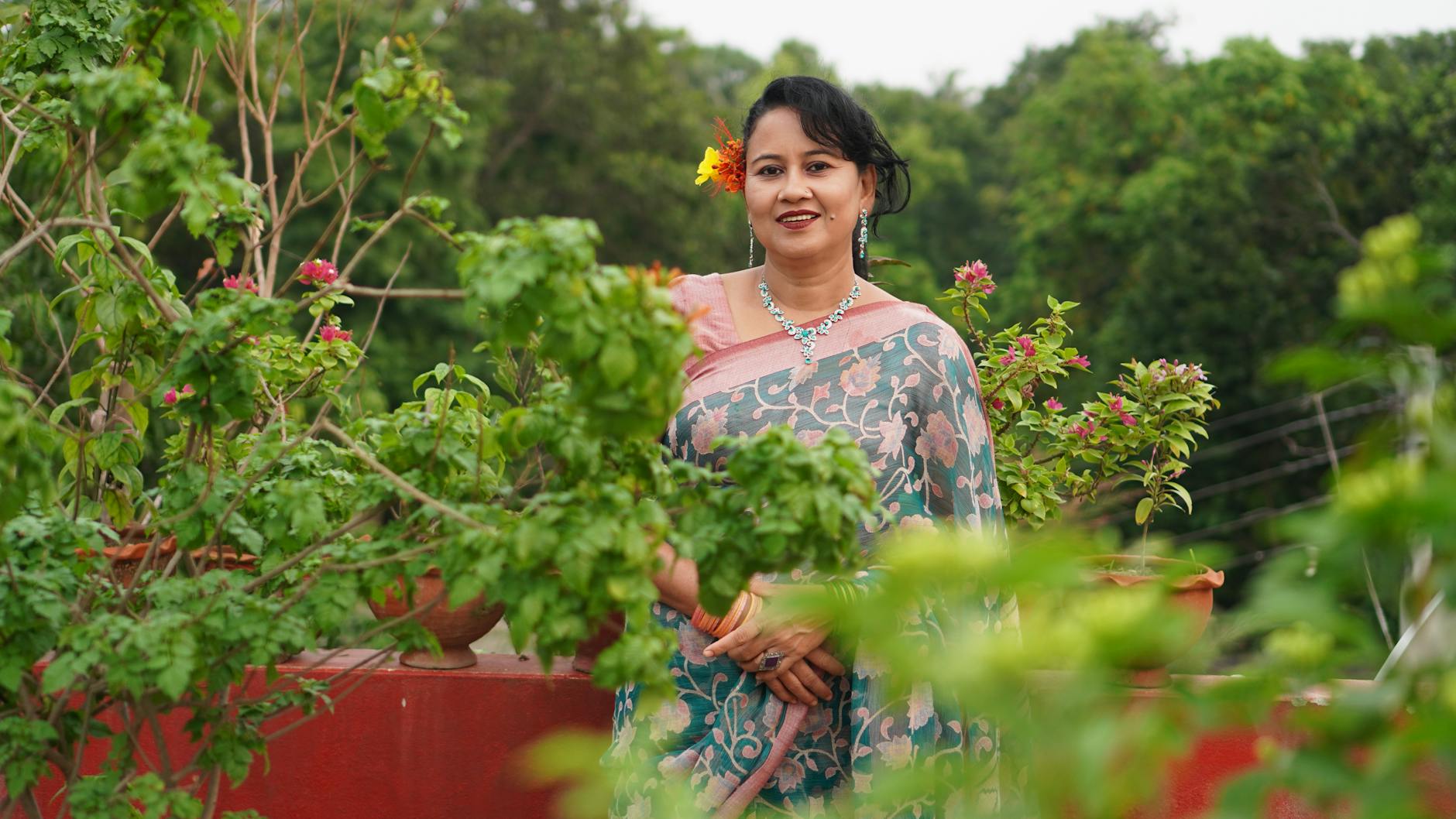 A woman in traditional sari poses outdoors among green plants, exuding grace and happiness.