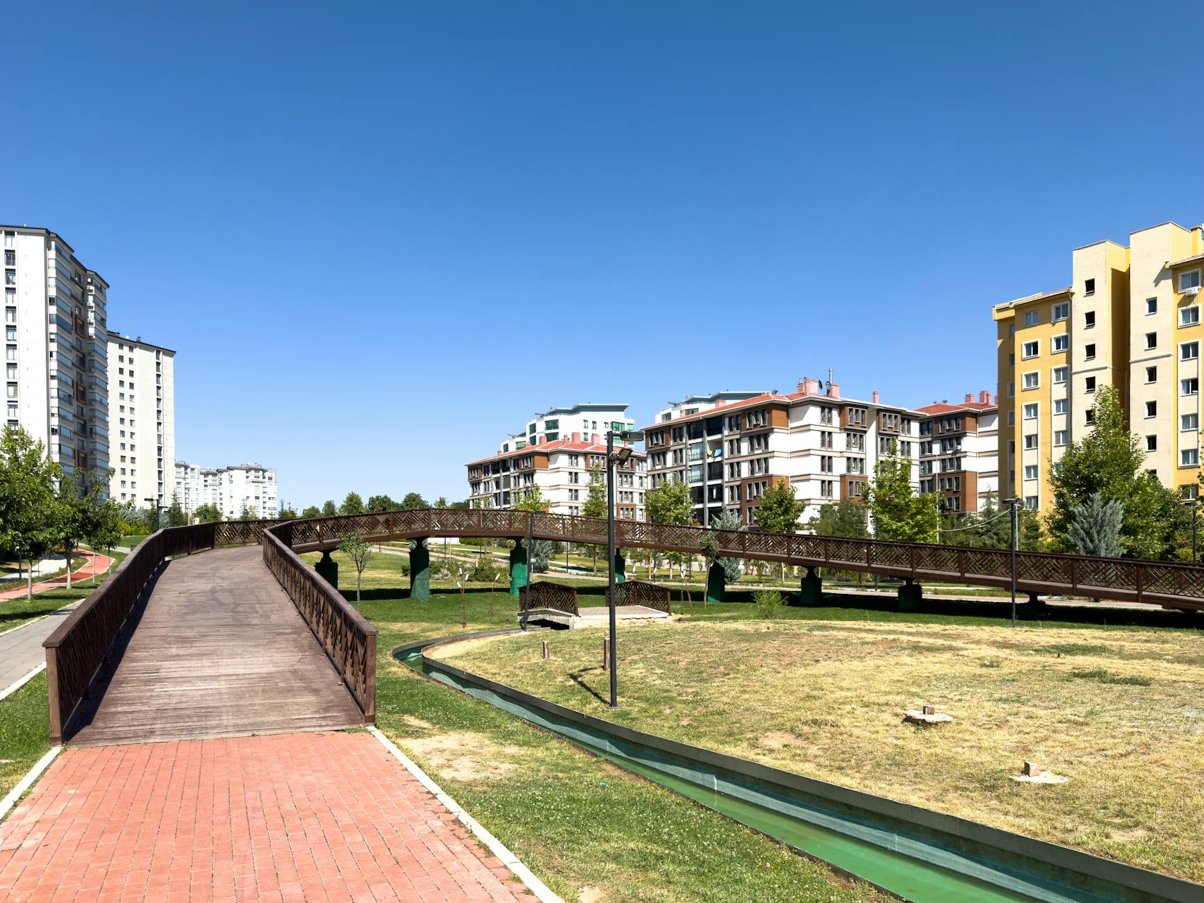 A sunny day in an urban park with modern buildings and a wooden bridge in Elâzığ, Türkiye.