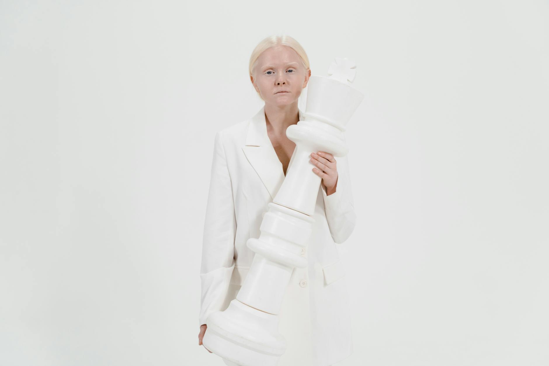Woman in white outfit holding a large white chess king against a plain background.