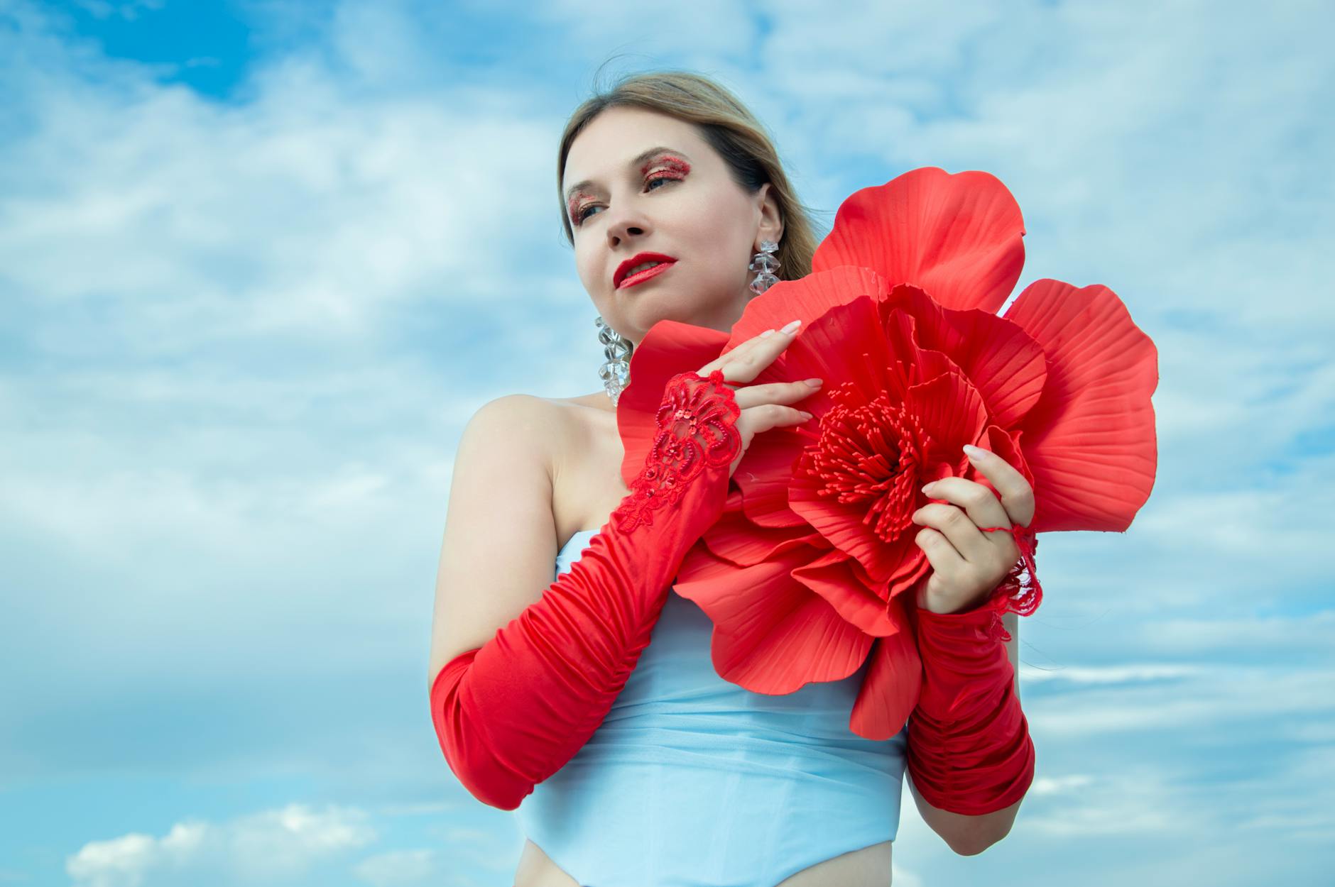 Stylish woman holding an oversized red flower against a clear blue sky, exuding fashion and elegance.