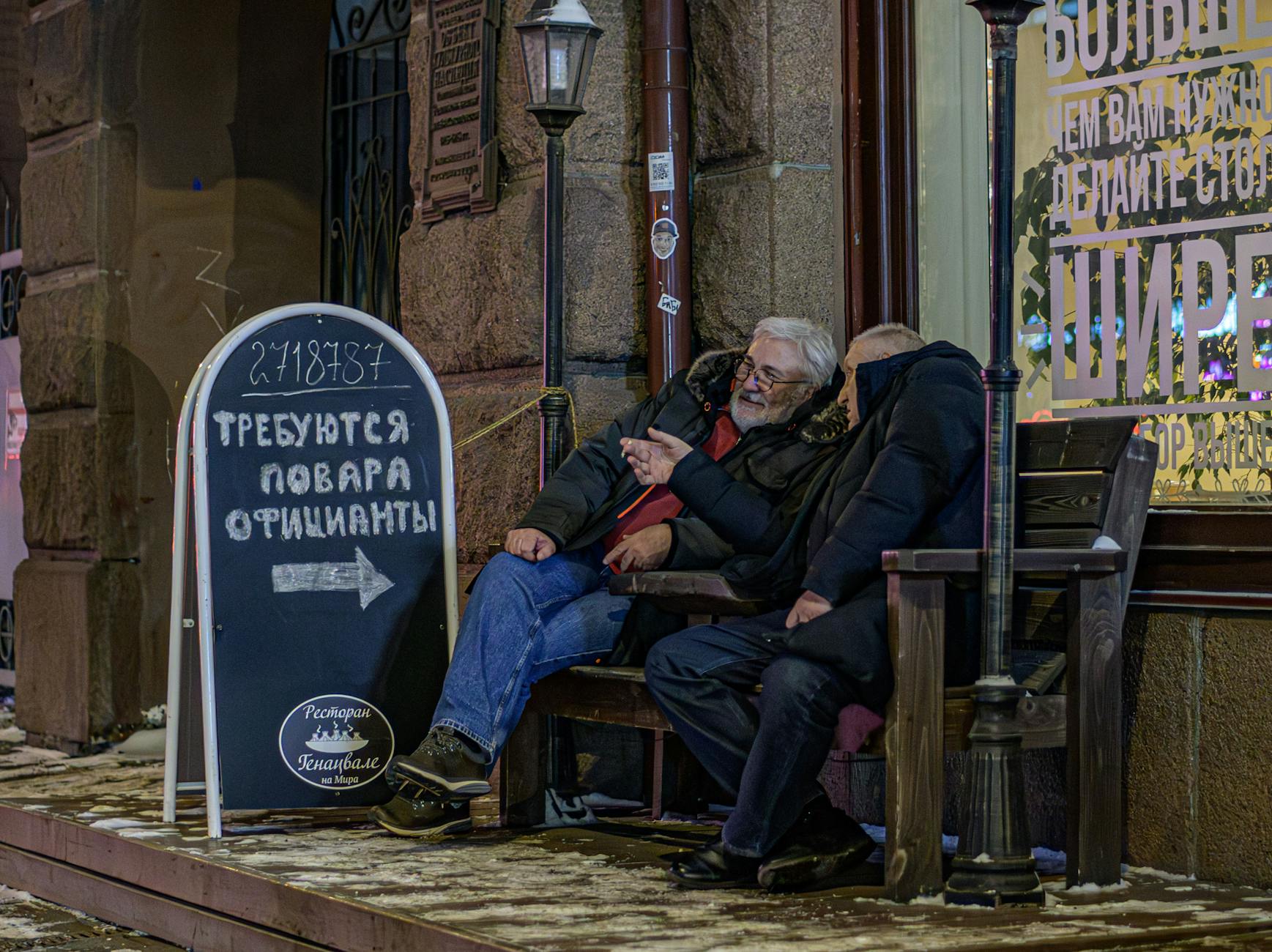 Two men relax outside a café on a winter night, reading a recruitment sign.