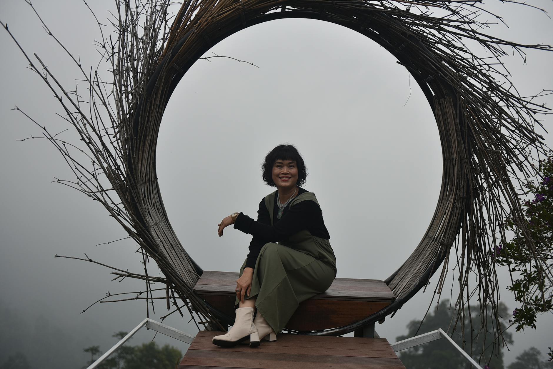 A woman smiling while seated in a large circular rustic frame with a misty backdrop.