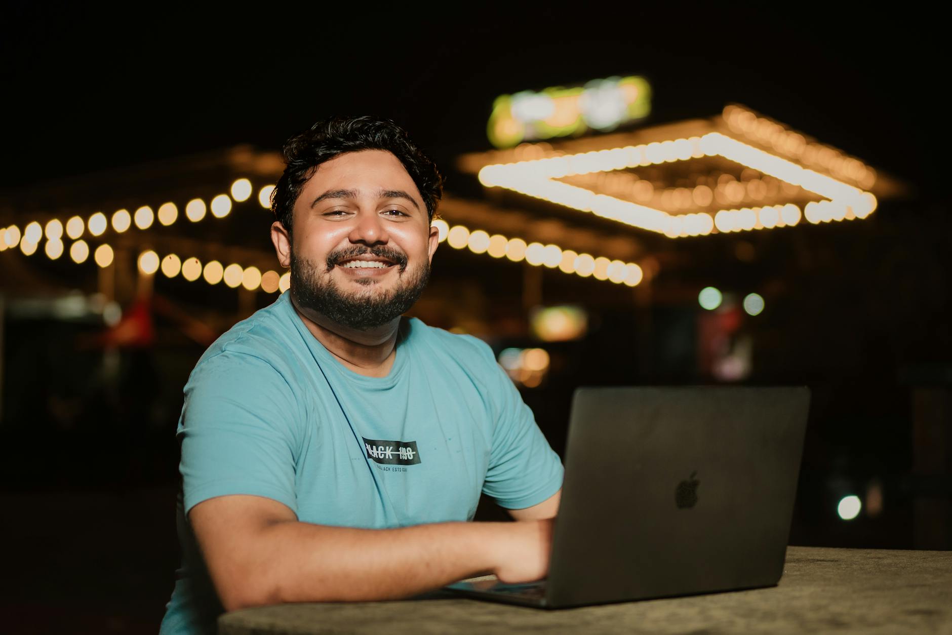 Happy man with beard and mustache sitting outdoors at night using a laptop, smiling warmly.