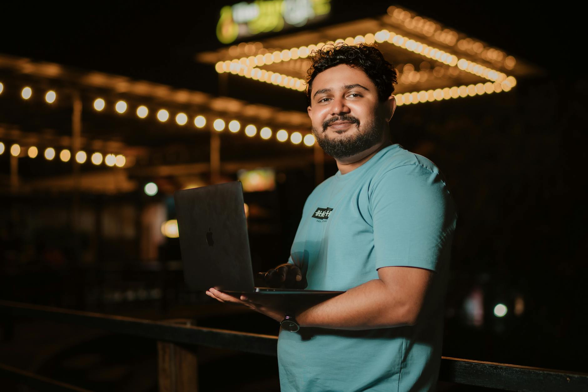 Man with a beard using a laptop outdoors at night, surrounded by string lights.