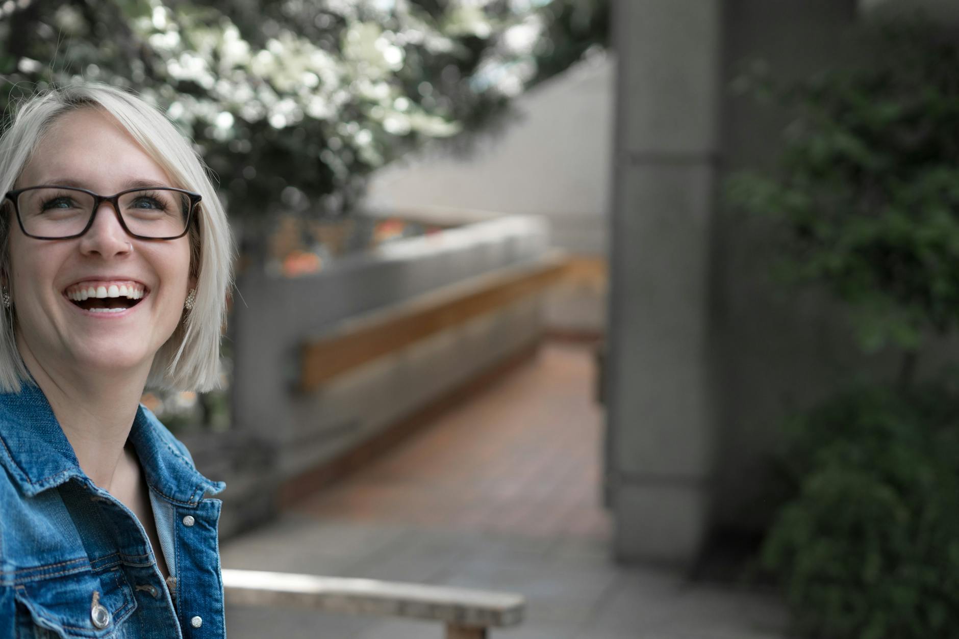 Joyful woman in glasses and denim jacket smiling in a serene outdoor setting.