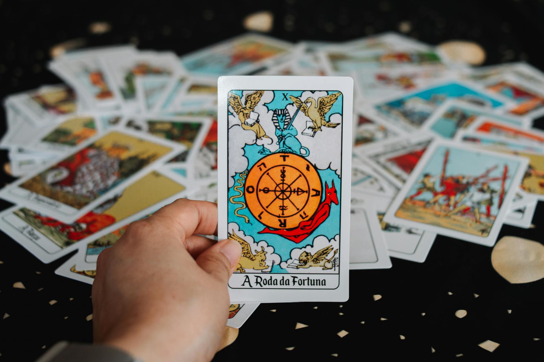 A hand holding 'The Wheel of Fortune' tarot card surrounded by scattered tarot cards.
