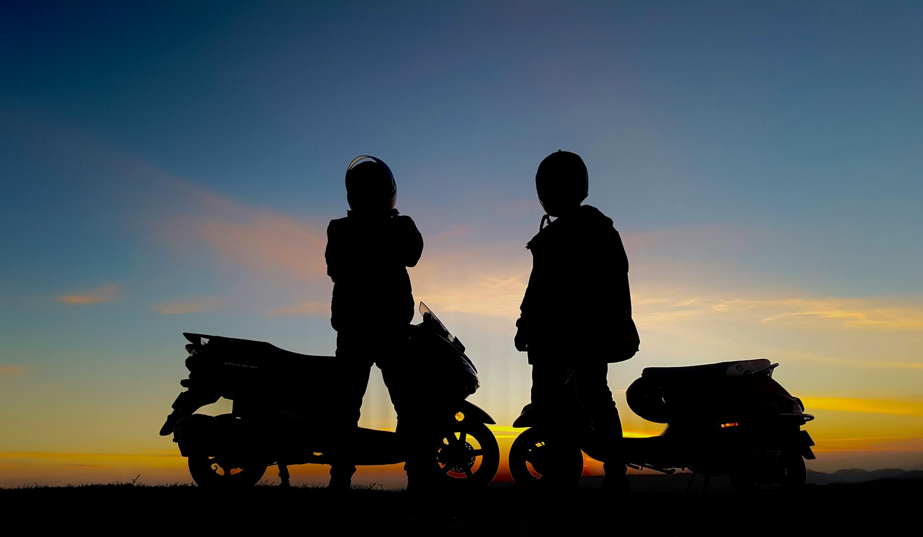 Silhouetted motorcyclists with scooters against a colorful sunset sky.