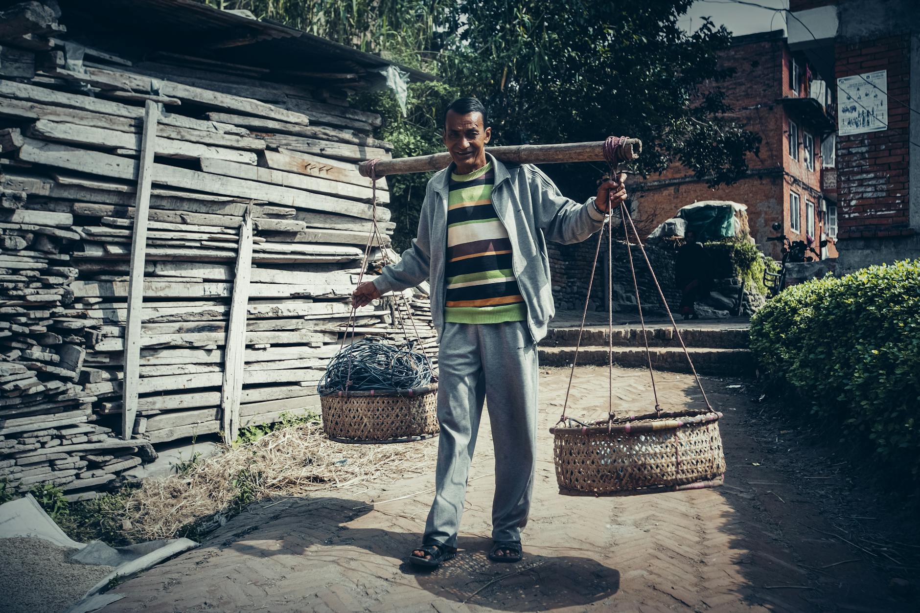 A smiling man in a striped shirt balances baskets on a pole, walking along a brick road bordered by wooden planks.