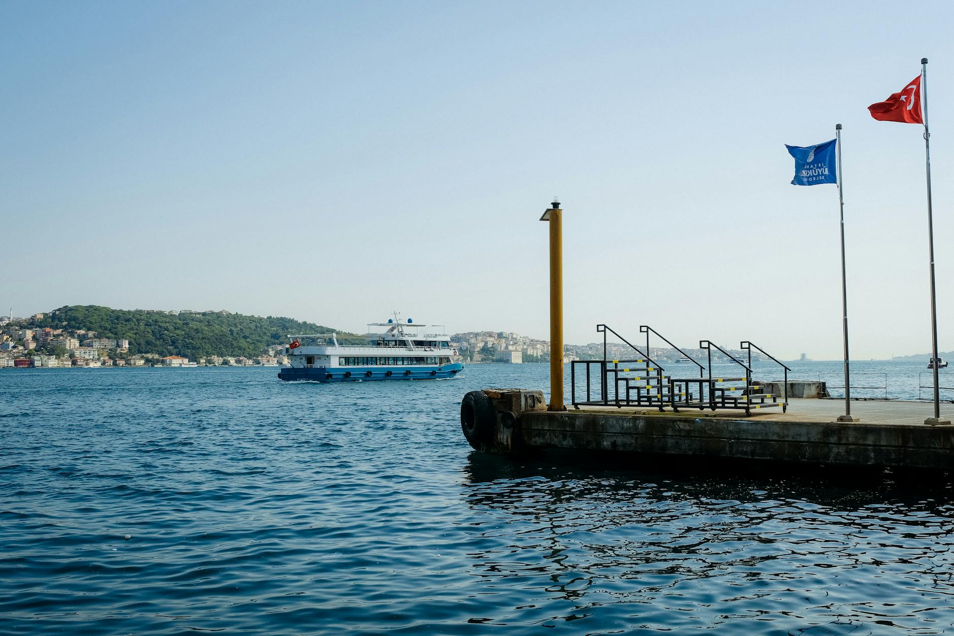 A ferry approaches a pier on Istanbul's vibrant waterway with cityscape views and vibrant flags.