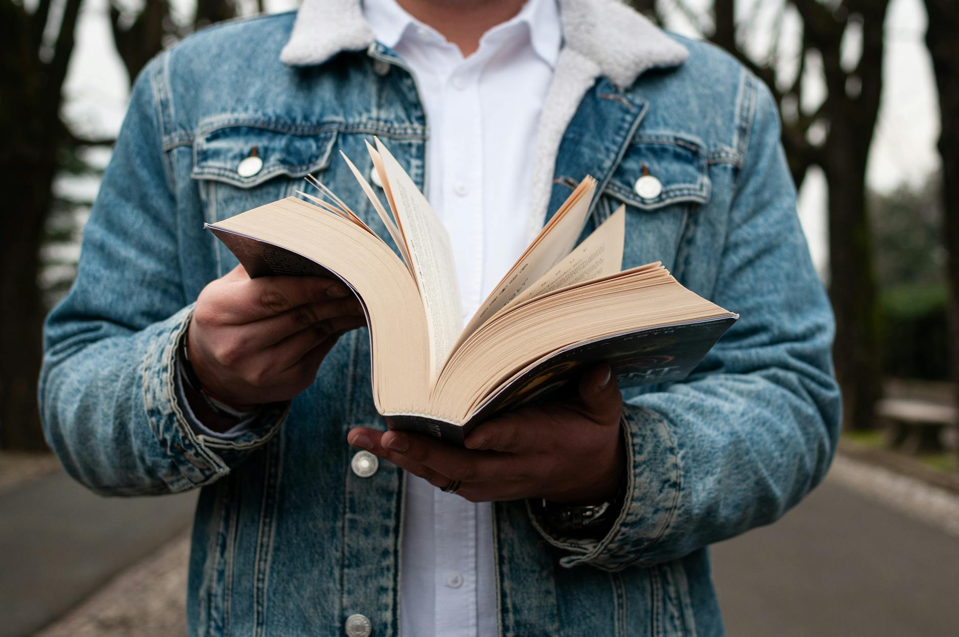 A person in denim jacket holding an open book in a Milan park. Calm and serene outdoor reading scene.