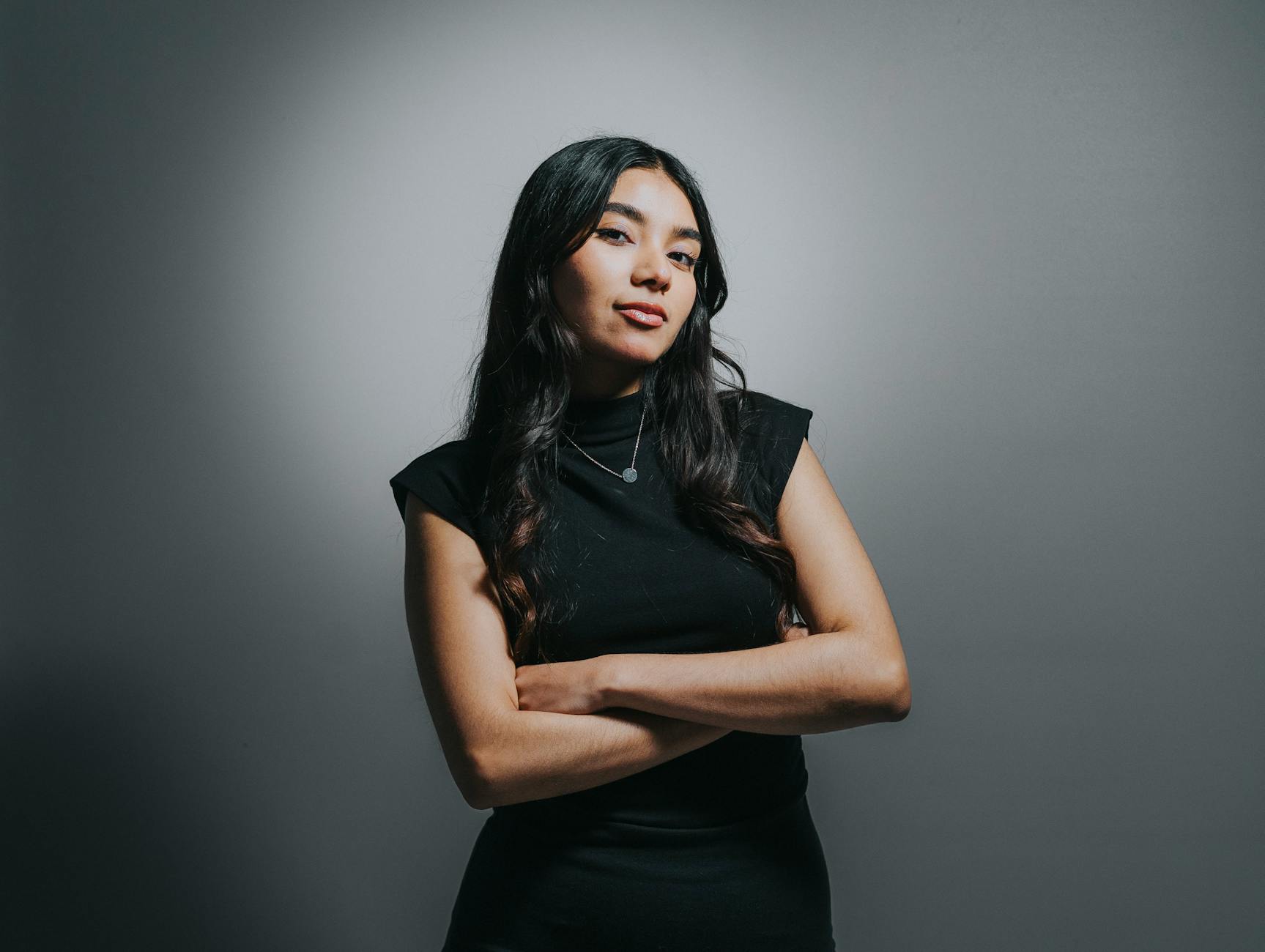 Portrait of a confident woman in a black dress, arms crossed, standing against a gray background.