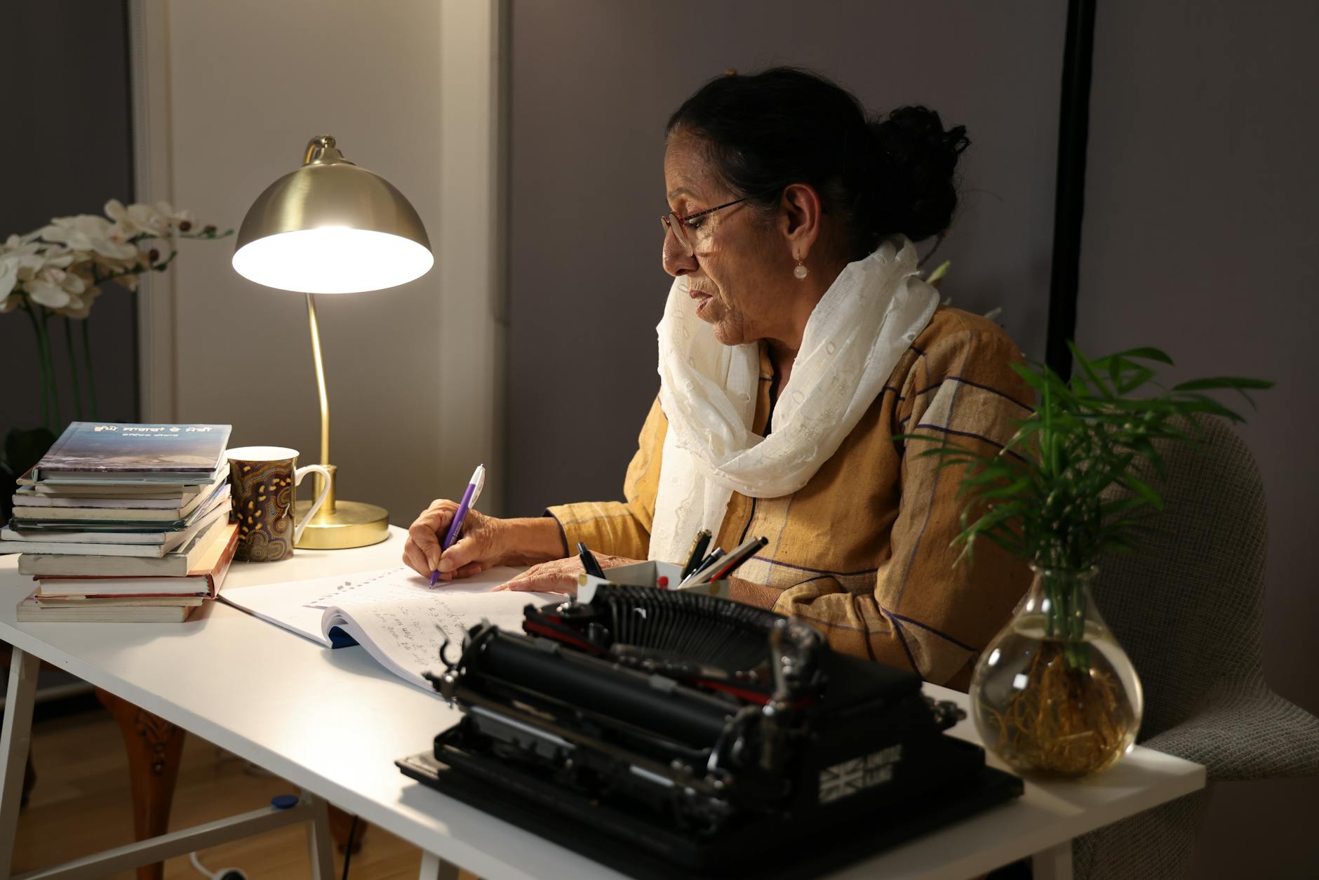 Senior woman writing at home desk with a typewriter and lamp.