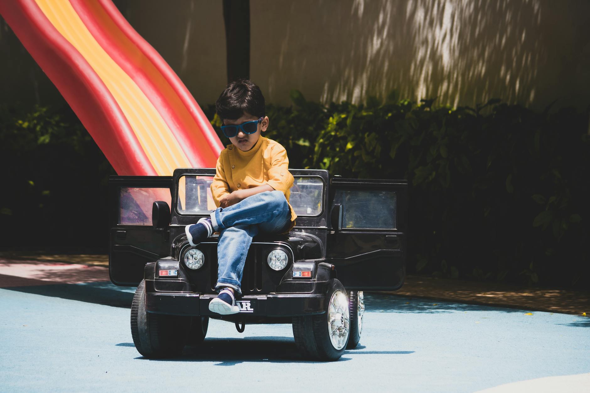 A young boy in sunglasses sits confidently on a toy car at an outdoor playground, enjoying a sunny day.