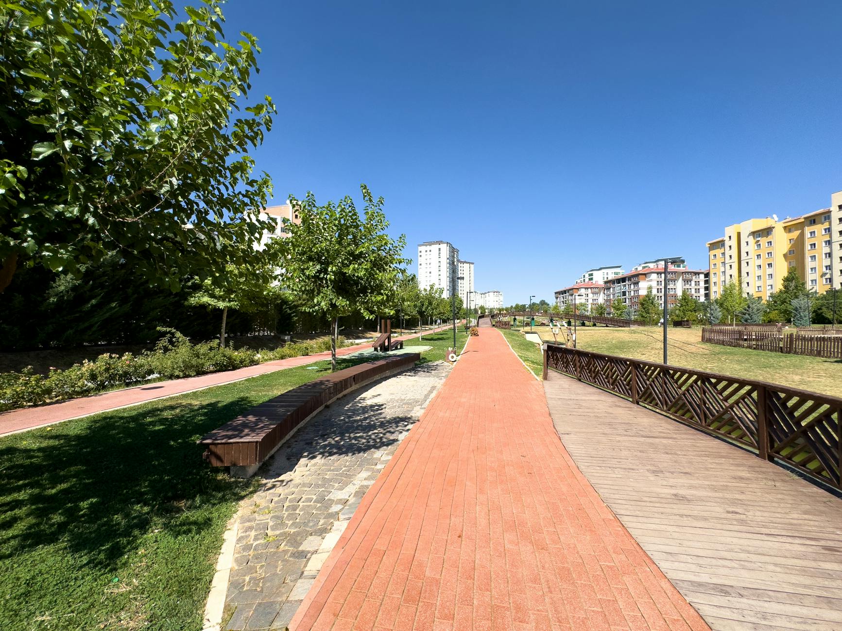 A vibrant, sunny park walkway with trees and buildings in Elâzığ, Türkiye.