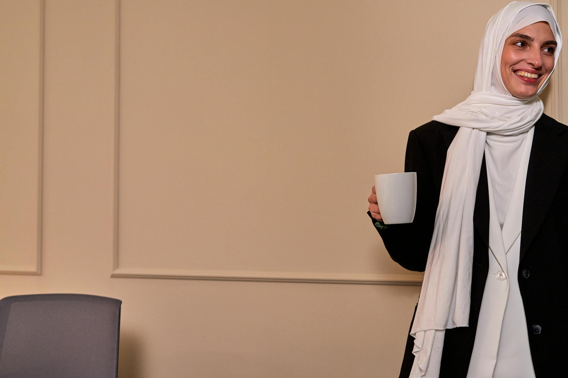 A cheerful Muslim woman in hijab holding a coffee cup and smiling in an office environment.