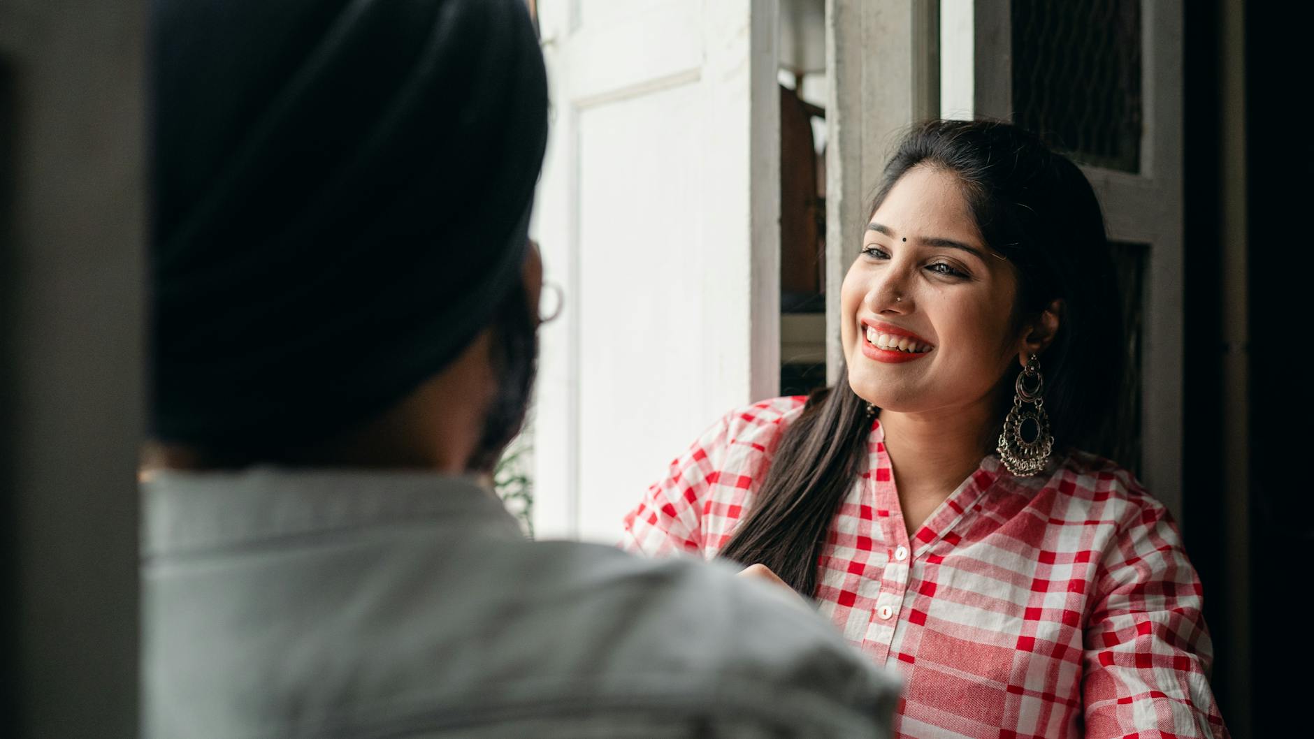Positive young Indian woman in causal shirt with traditional earrings communicating with anonymous blurred male and laughing candidly at funny story while both sitting in doorway