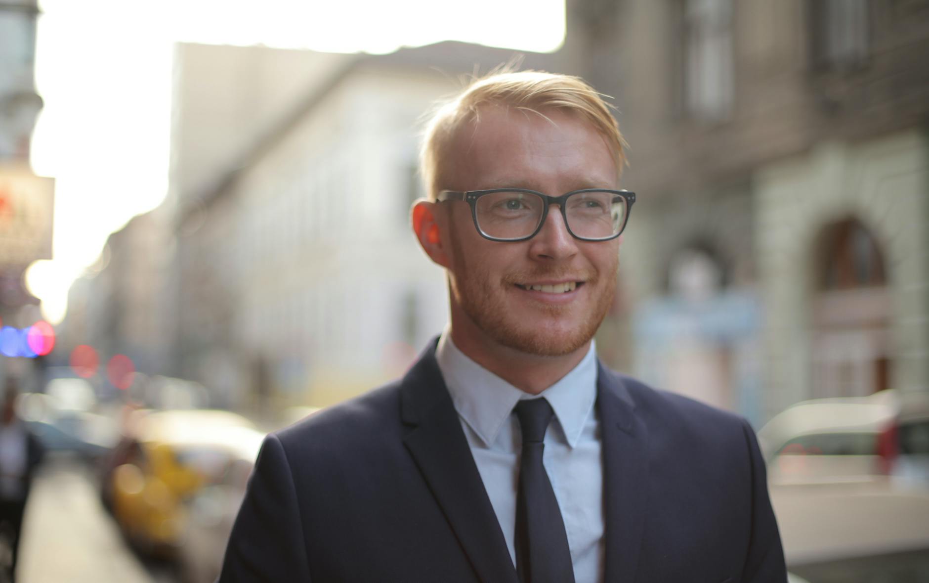 Smiling bearded male employee wearing formal clothes and eyeglasses looking away while standing on street against city downtown