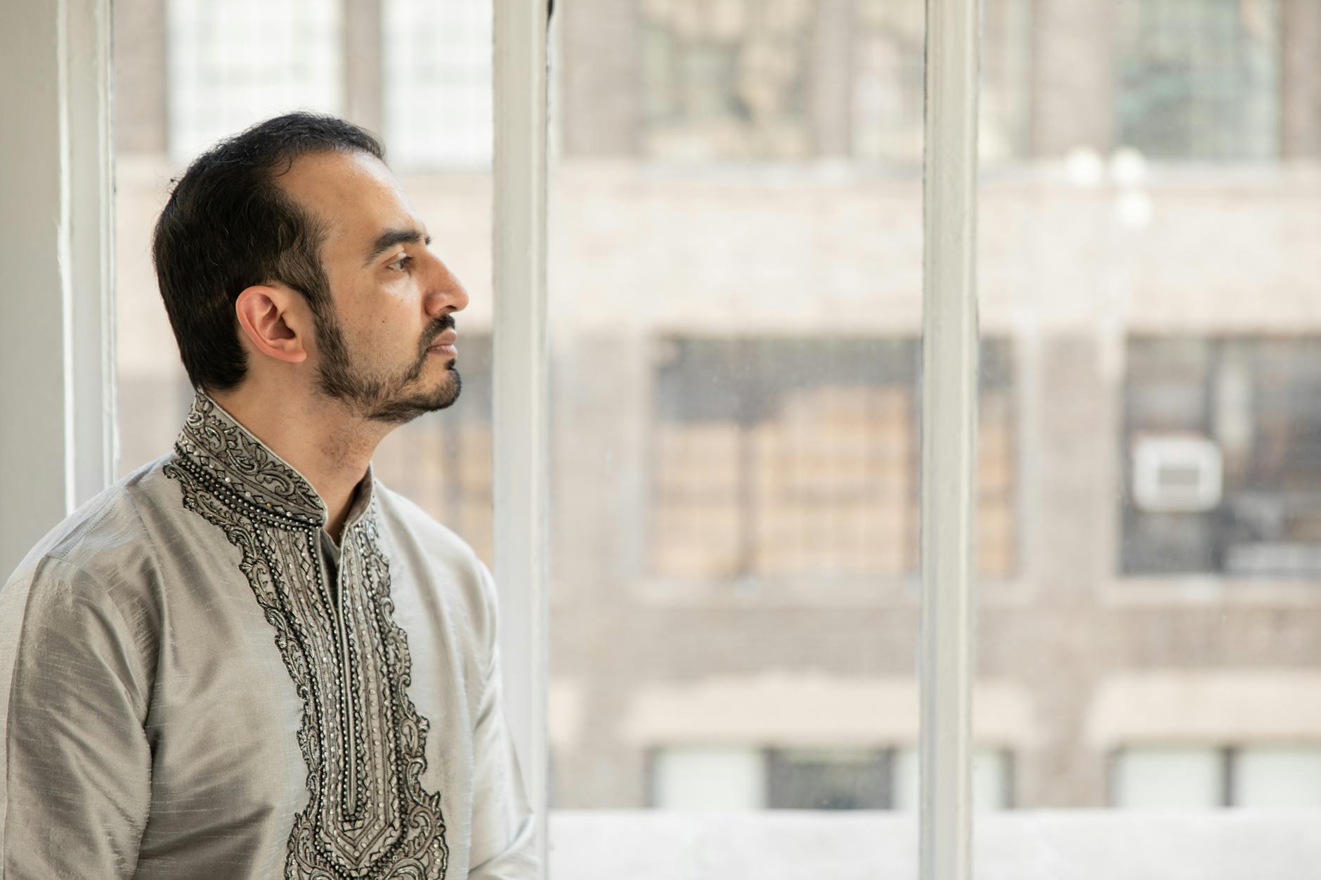 A man in traditional attire gazes thoughtfully out a city window, capturing a serene moment.