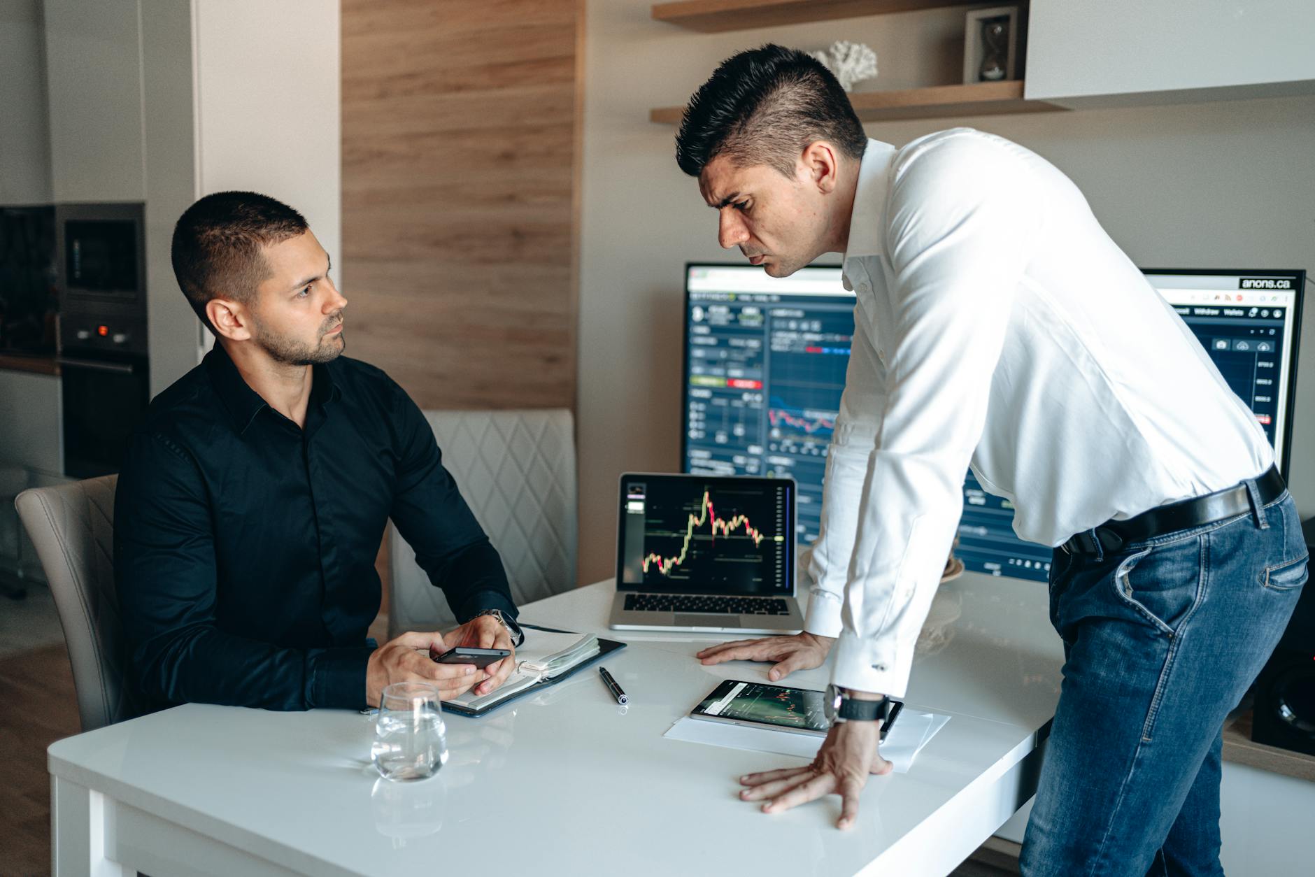 Two businessmen analyzing stock market data on laptops and tablets in an office environment.