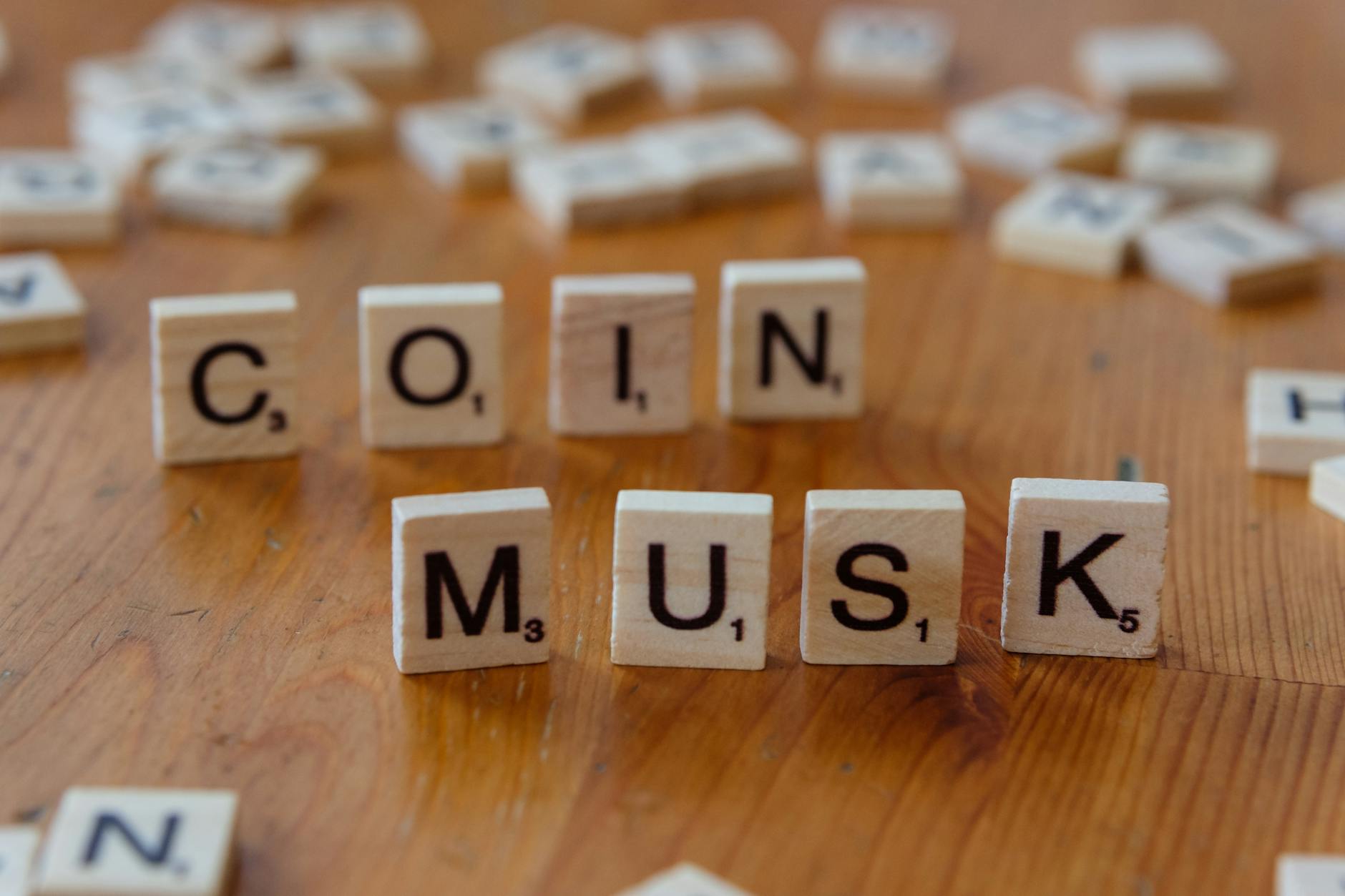 Scrabble tiles forming the words 'COIN' and 'MUSK' on a wooden table surface.