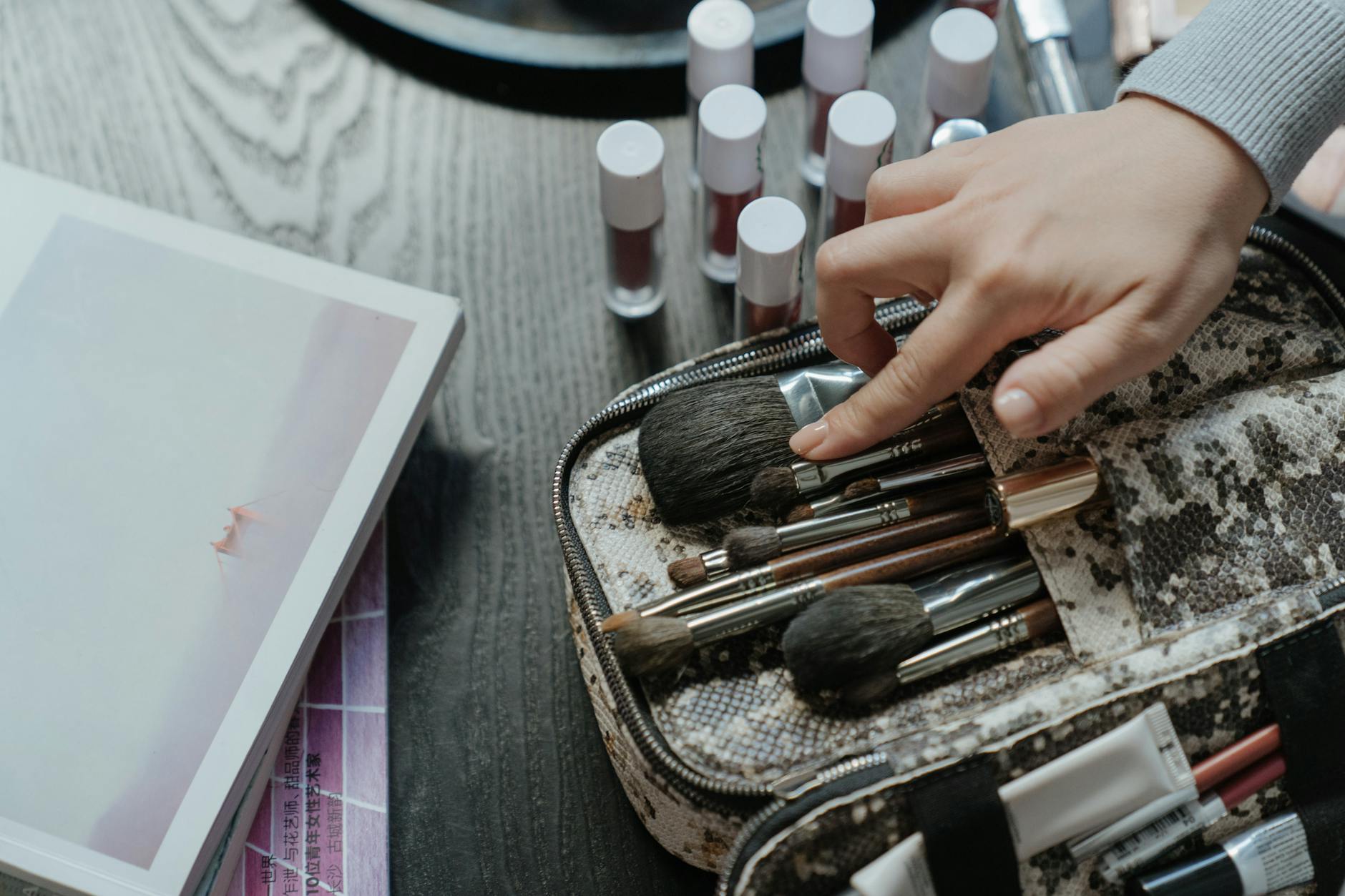 A hand arranging makeup brushes in a case surrounded by cosmetics in a flat lay setup.