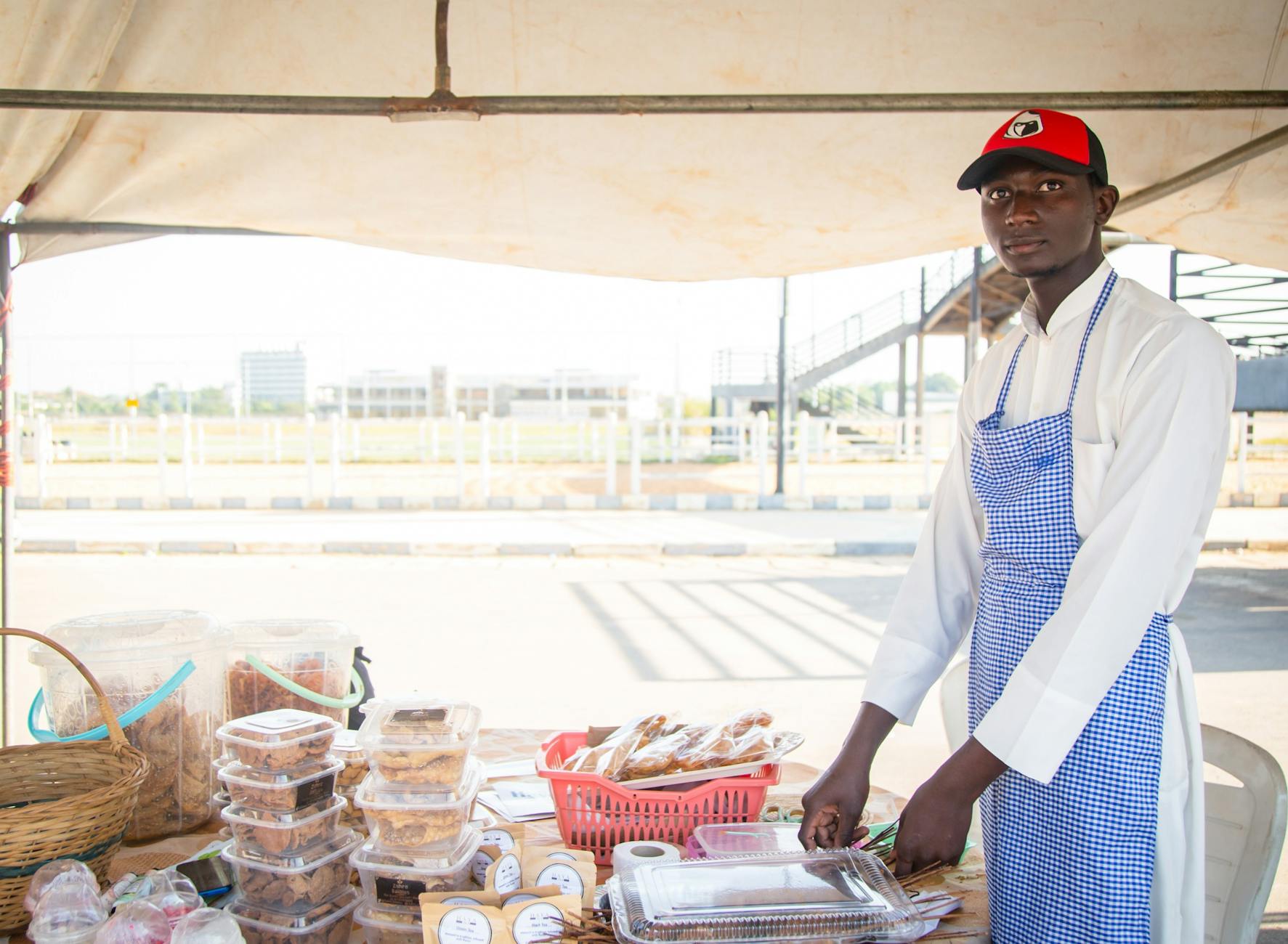 A young male vendor in Kaduna, Nigeria, selling baked goods at an outdoor market stall.