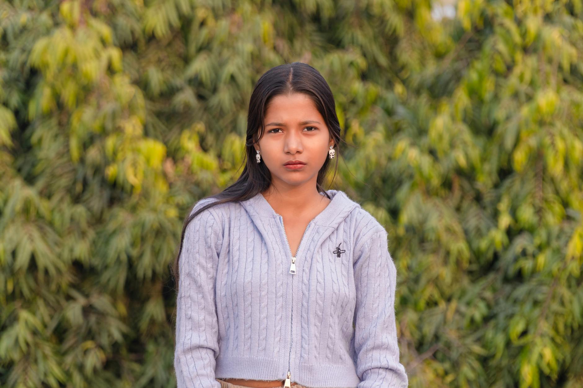 Portrait of a serious teenage girl in a cable knit sweater, standing outdoors with a green foliage background.