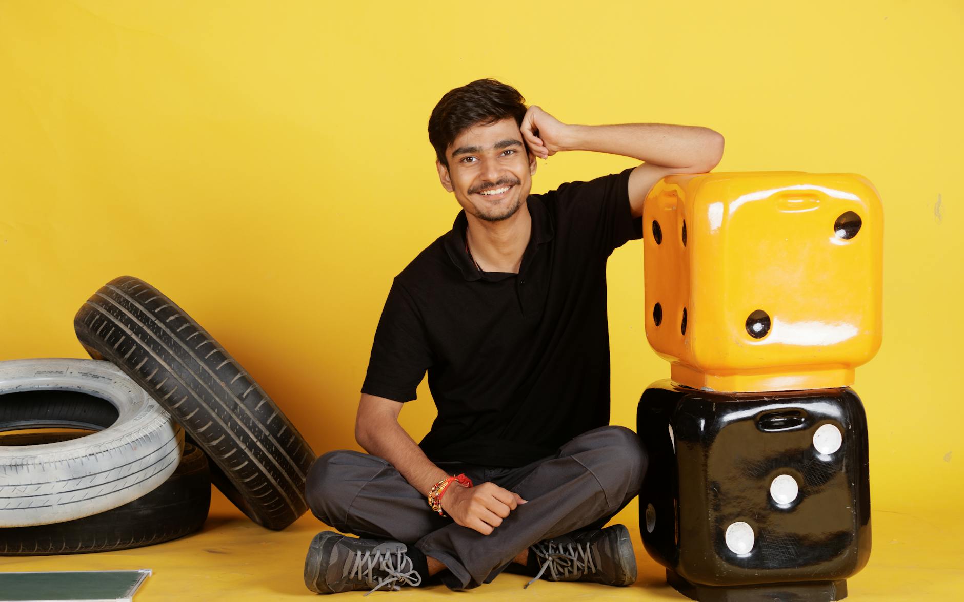 Smiling young man seated indoors with large dice and tires, exuding a positive and relaxed vibe.