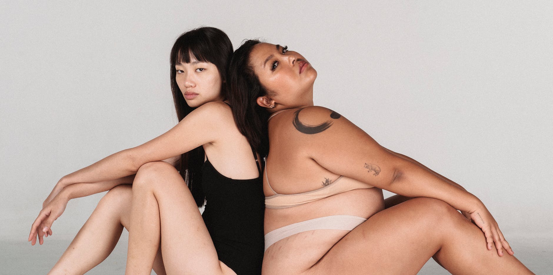 Asian female models with different body constitution wearing underwear and looking at camera while sitting on white background back to back