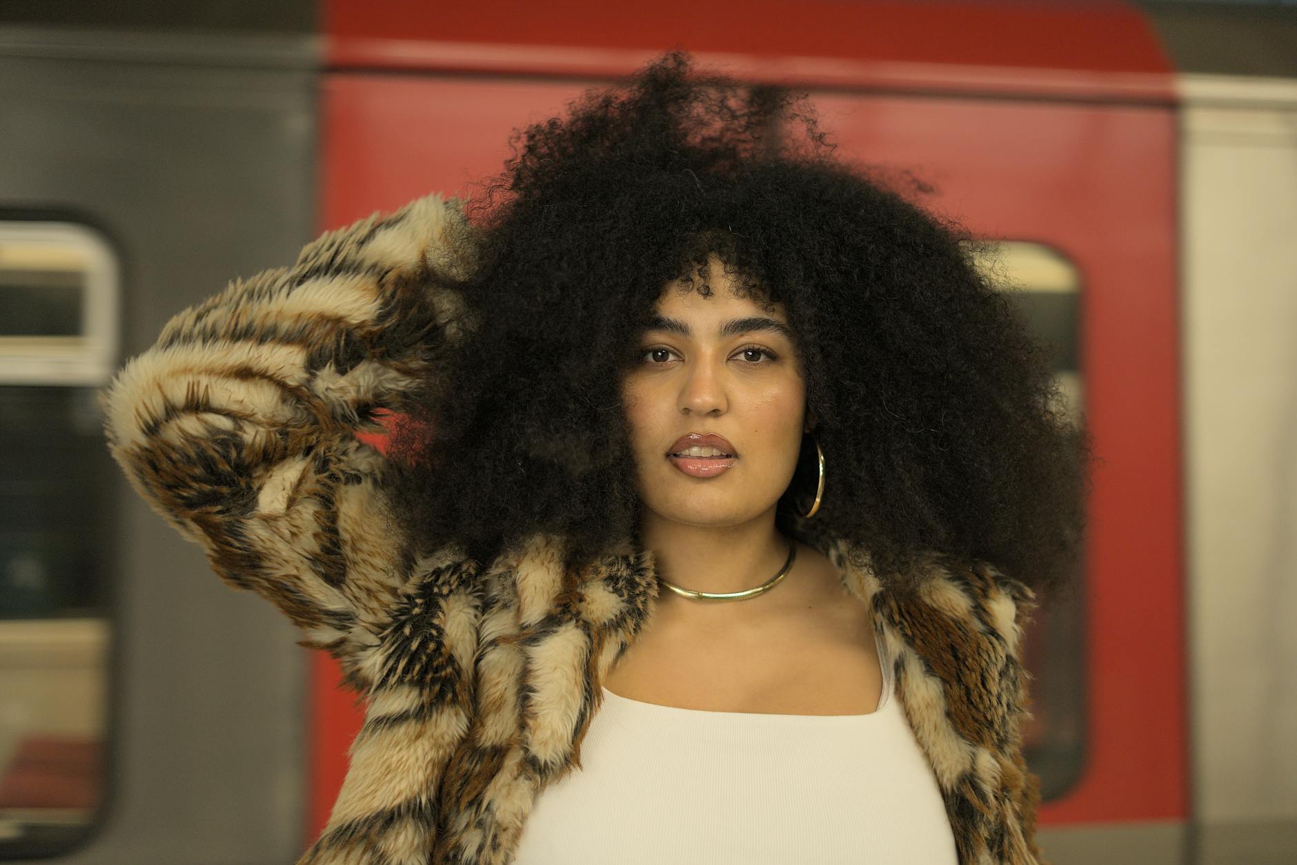 Portrait of a stylish woman with an afro, wearing a fur coat, posing confidently in front of a moving subway train.