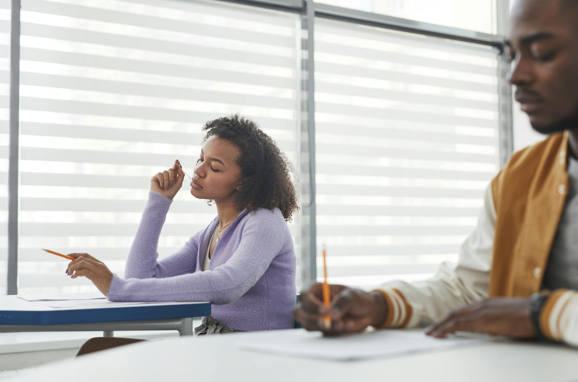 Two students in a classroom setting, focused on taking an exam with pencils in hand.
