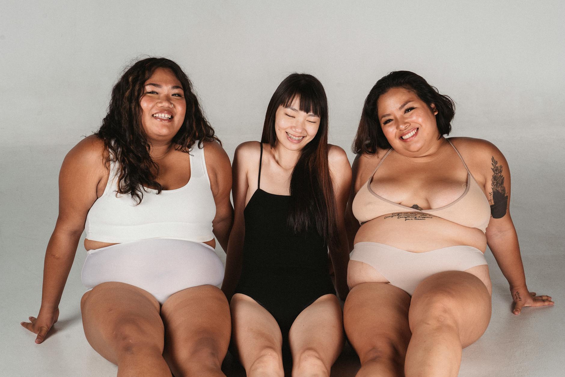 Positive slim Asian woman sitting on floor between cheerful overweight models in lingerie and looking at camera on white background