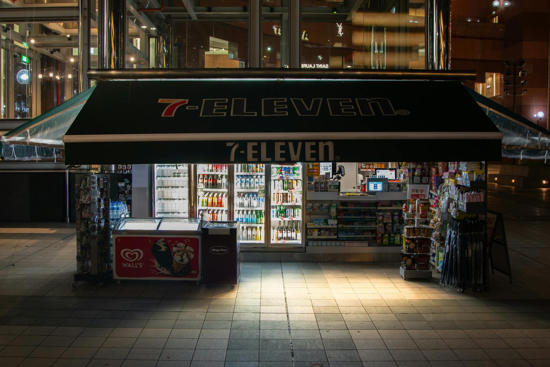 A brightly lit 7-Eleven convenience store at night, showcasing drinks and snacks.