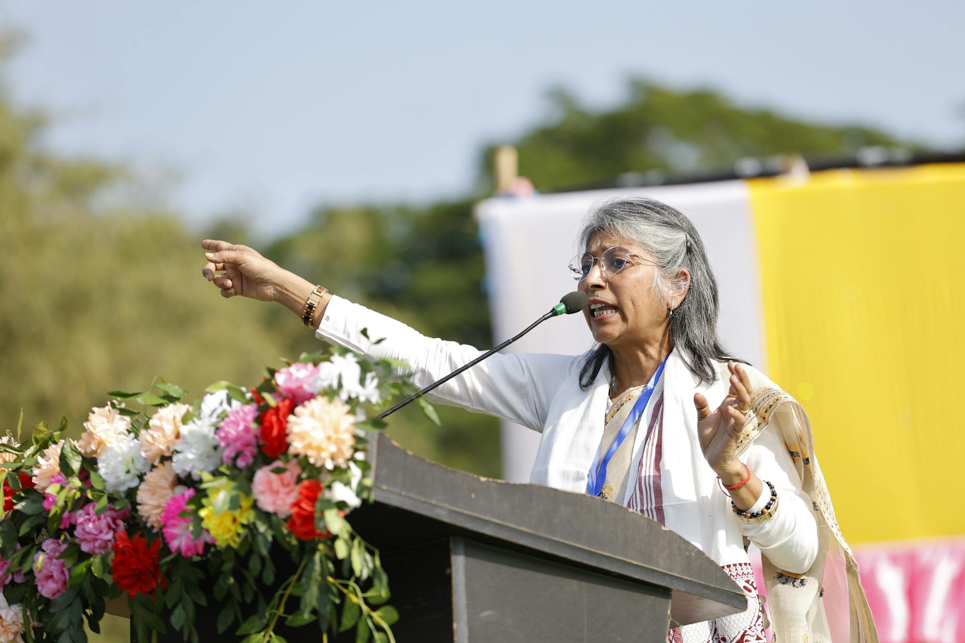 An Indian woman passionately speaks at an outdoor event with floral decorations.