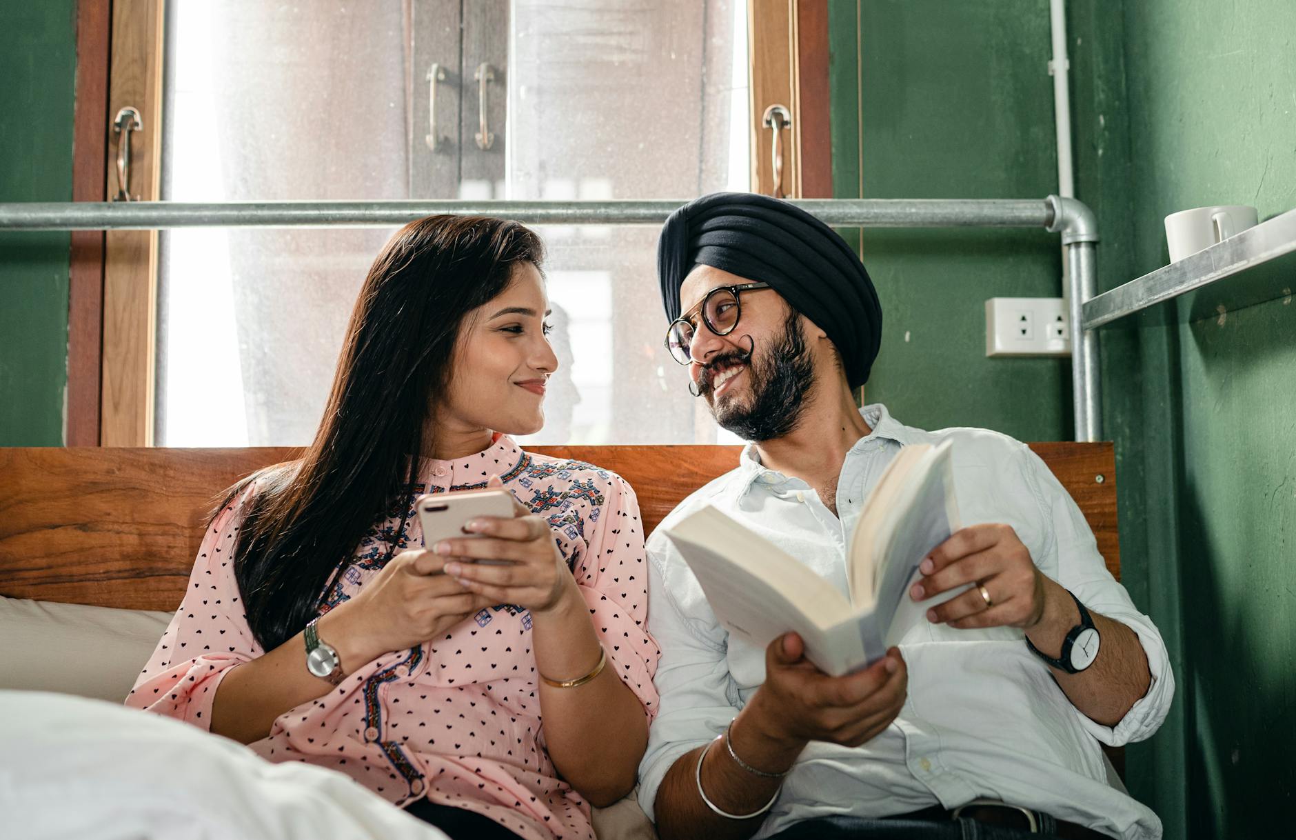 Optimistic content young Indian couple wearing casual clothes resting on bed with book and modern smartphone while smiling and looking at each other