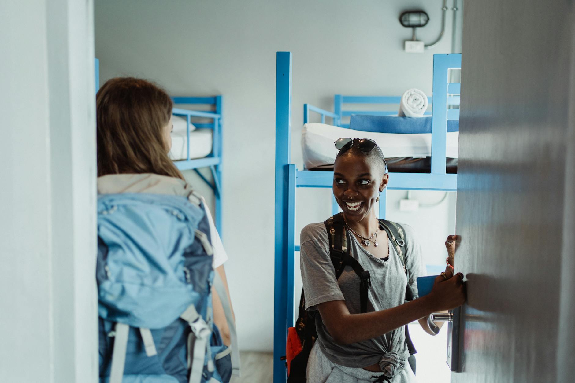 Two women backpackers entering a hostel dorm room with bunk beds, smiling and excited for their adventure.