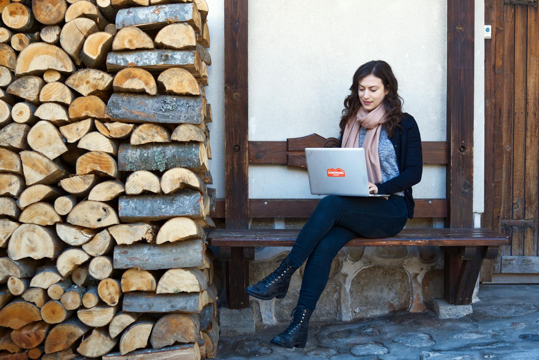 A woman sits on a wooden bench working on a laptop beside a stacked pile of firewood.