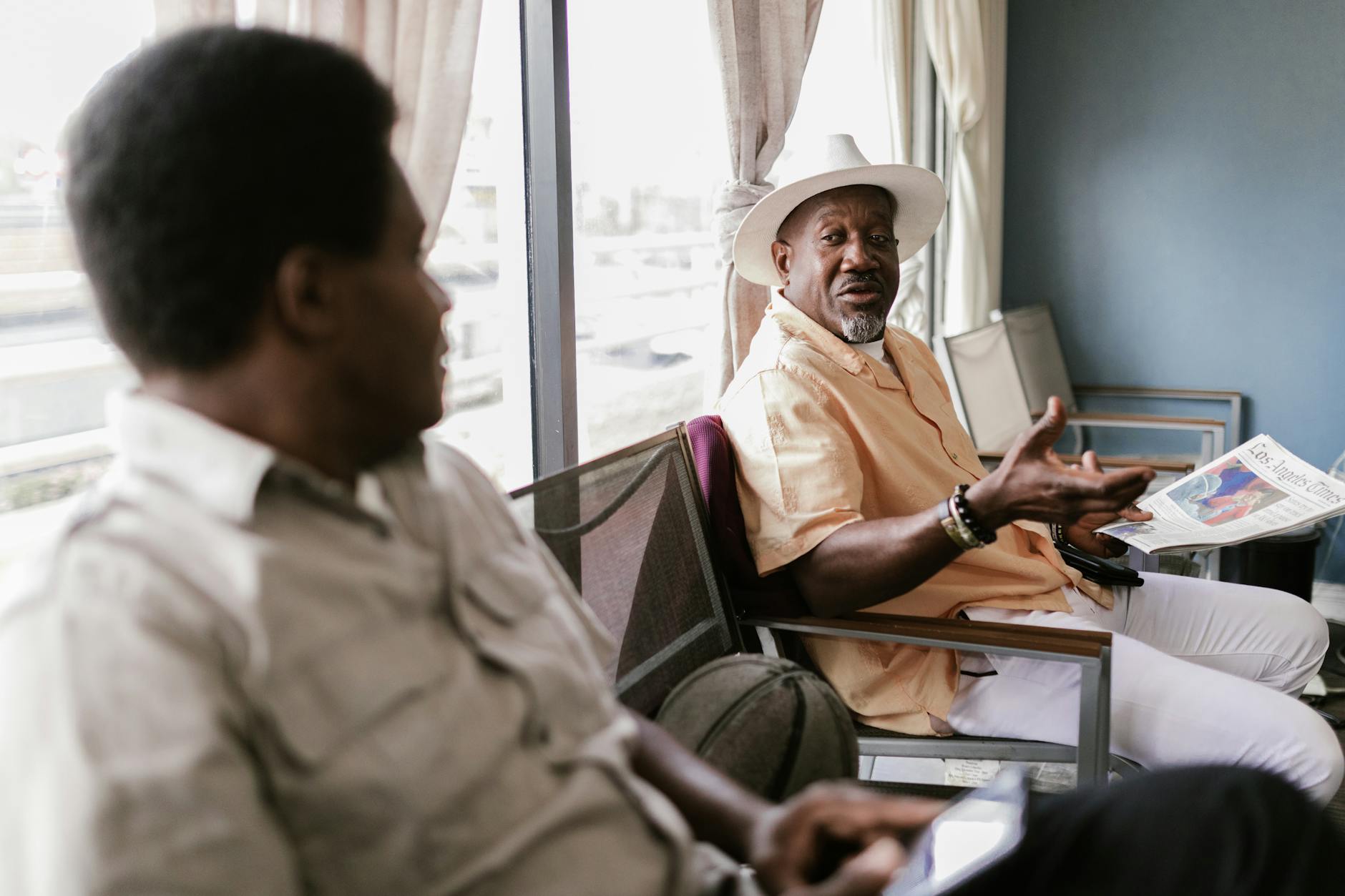 Two mature men engage in conversation in a comfortable indoor setting, one holding a newspaper.