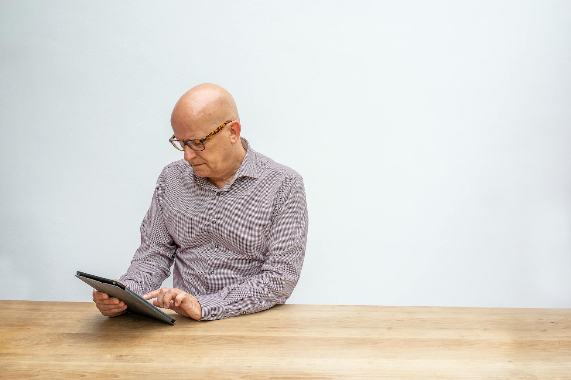 Elderly man in glasses using a tablet on a wooden table against a white background.