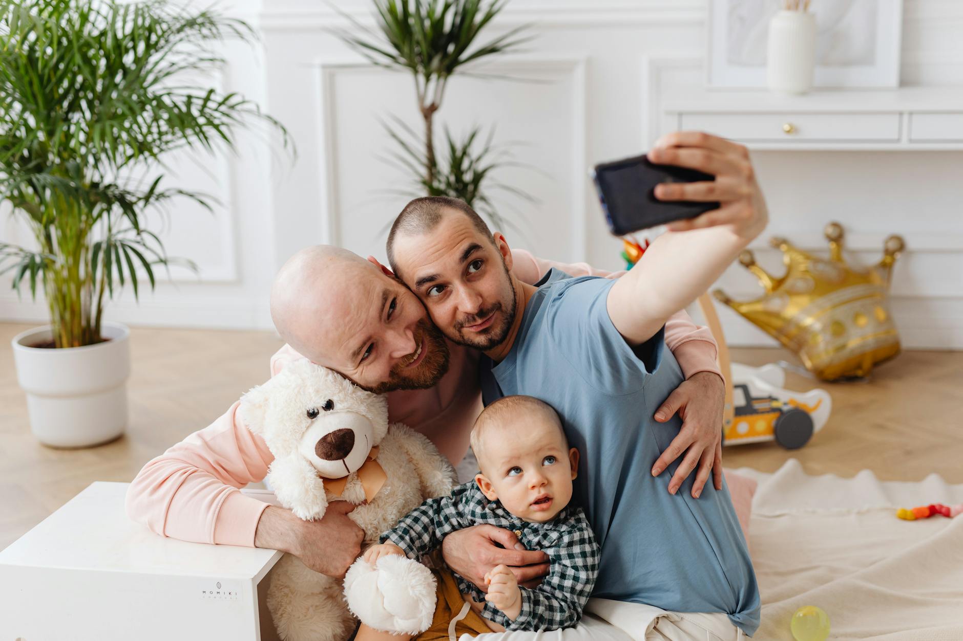 Two fathers and their toddler son taking a selfie in a cozy indoor setting.