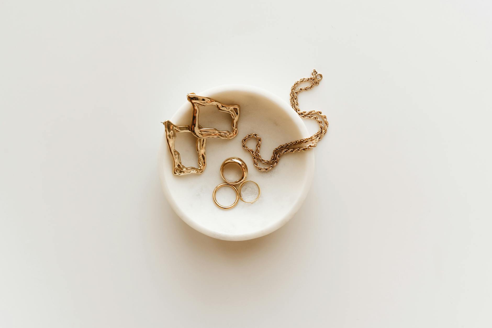 Stylish arrangement of gold jewelry including rings, earrings, and a necklace on a marble dish.
