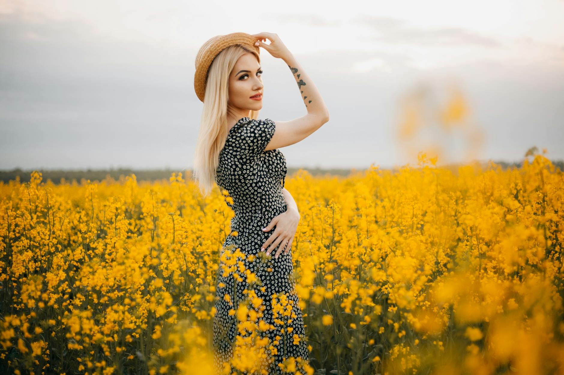 Blonde woman in a sundress and hat poses in a meadow of yellow flowers, embodying summer vibes.