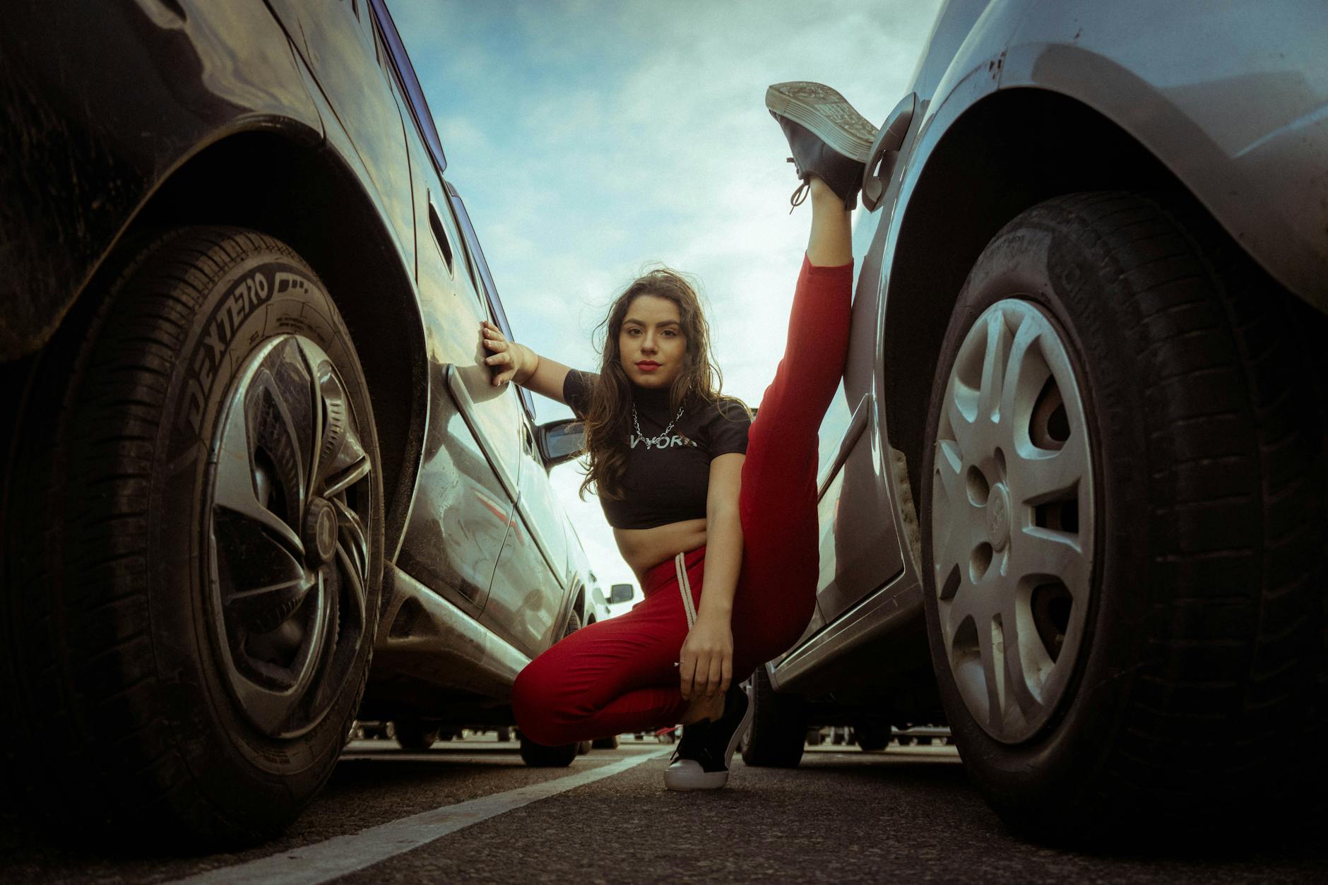 A confident woman poses with a high kick between parked cars under a cloudy sky.