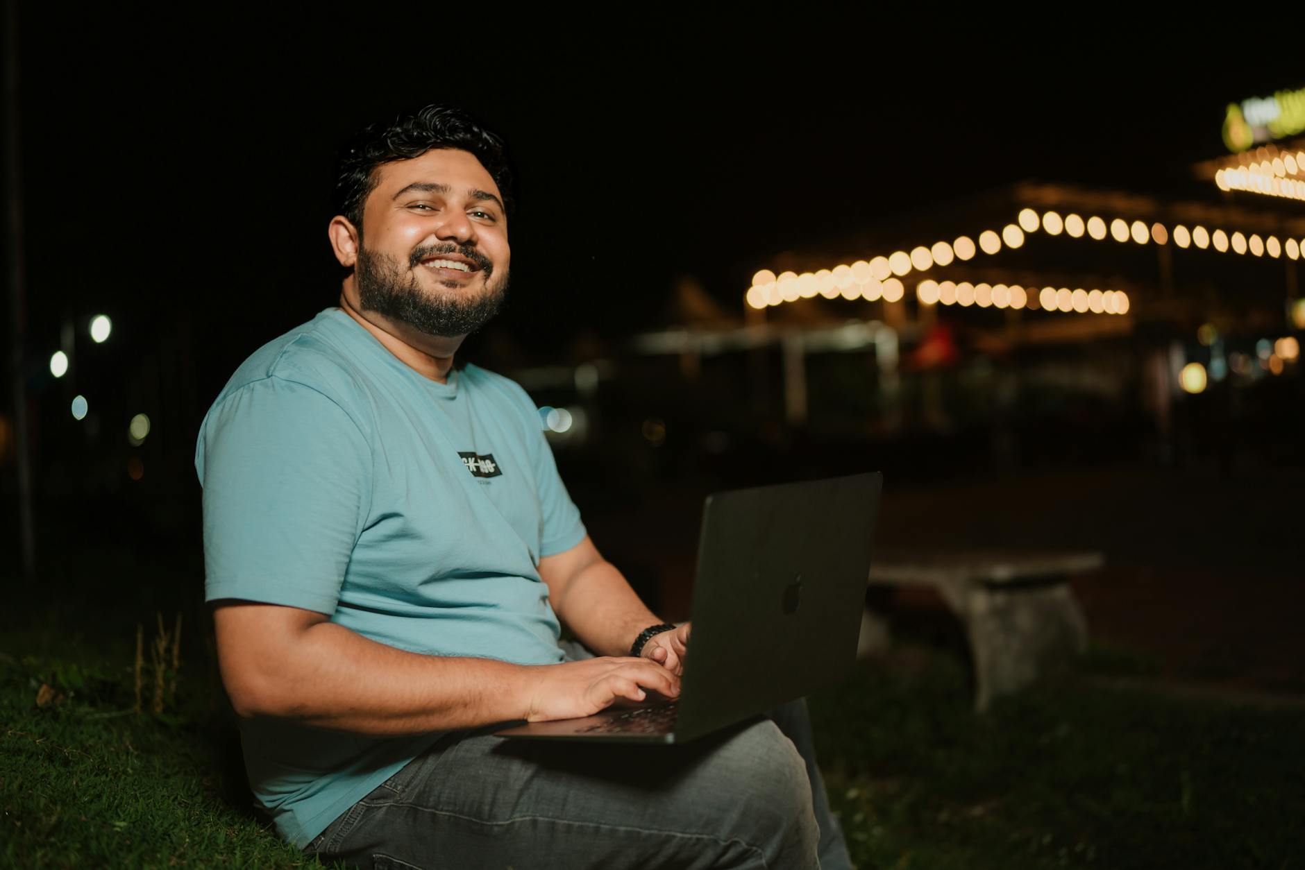 Cheerful man with laptop sitting outdoors at night, illuminated by warm lights.
