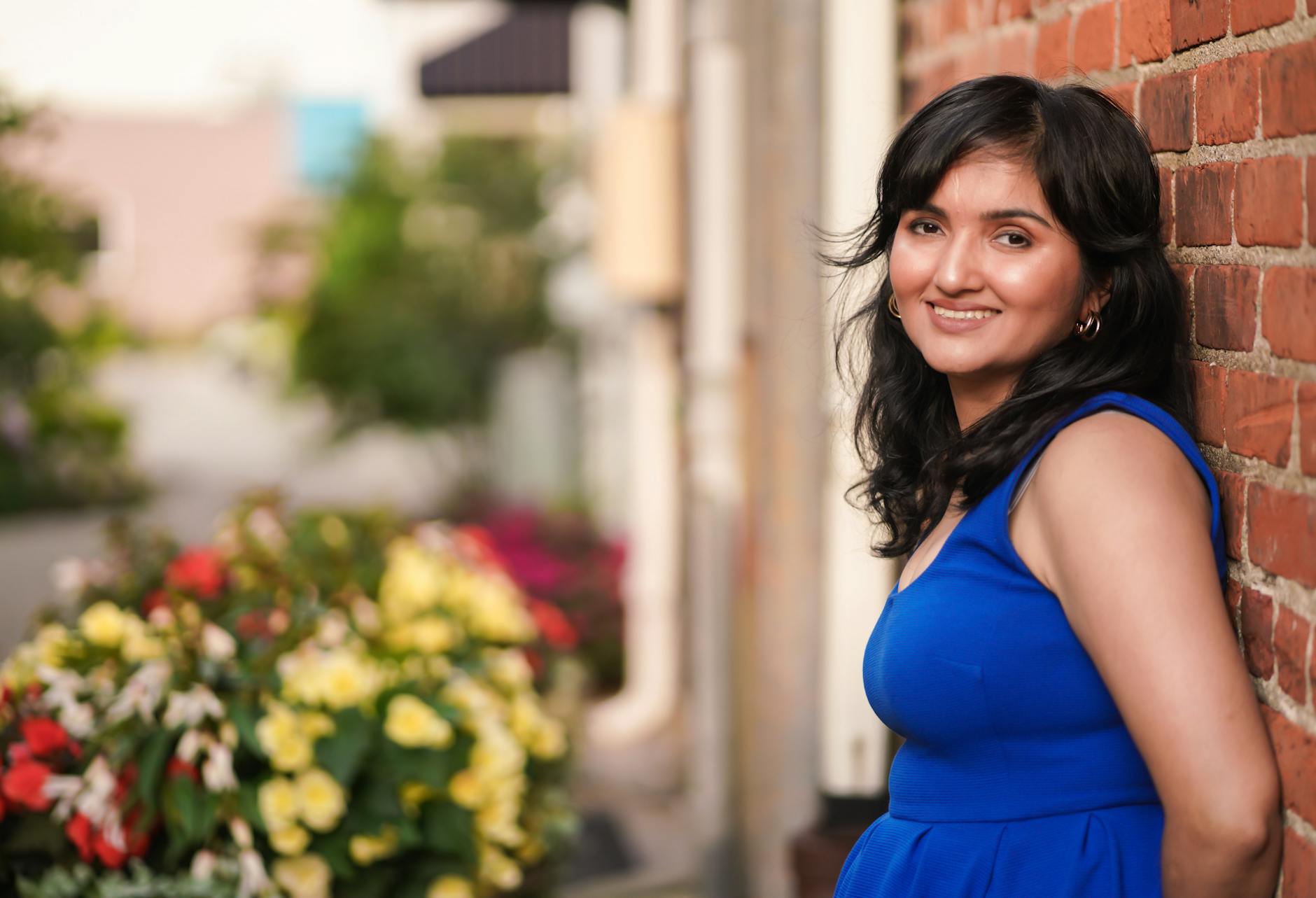 Smiling woman in a blue dress leaning against a brick wall with blurred flowers.
