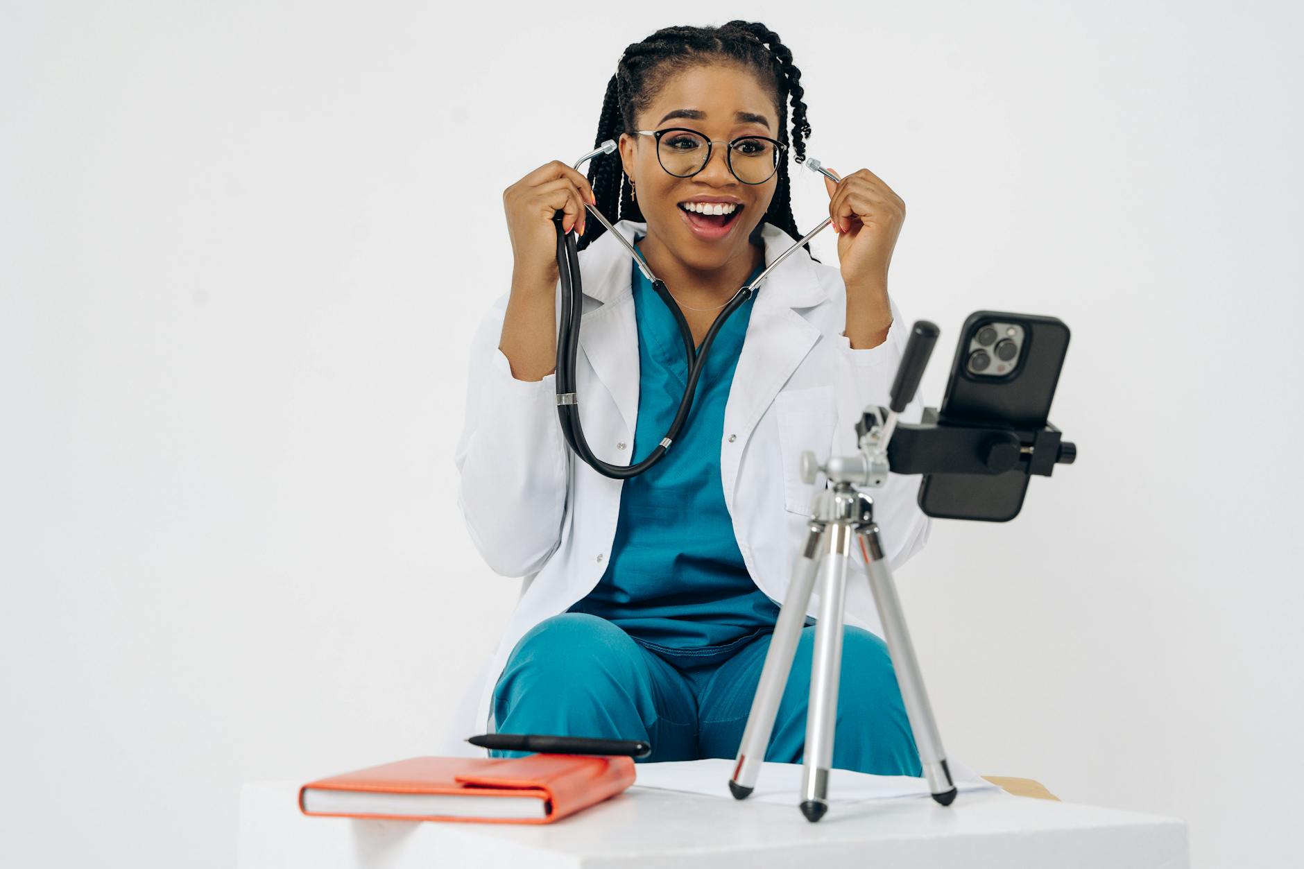 A cheerful female doctor in scrubs and white coat using a smartphone for a virtual medical consultation.