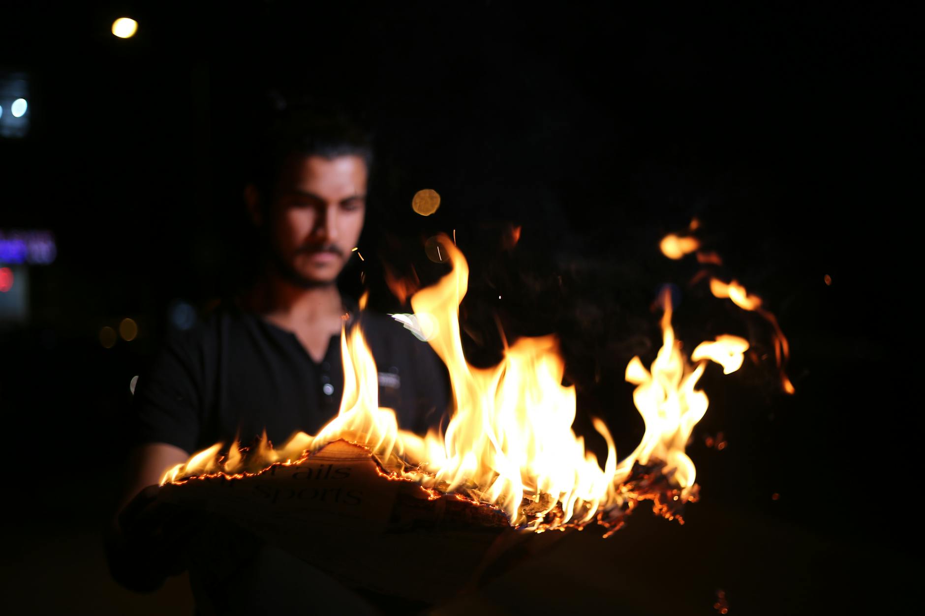 A man in a black shirt holds a flaming paper at night with a blurred background.