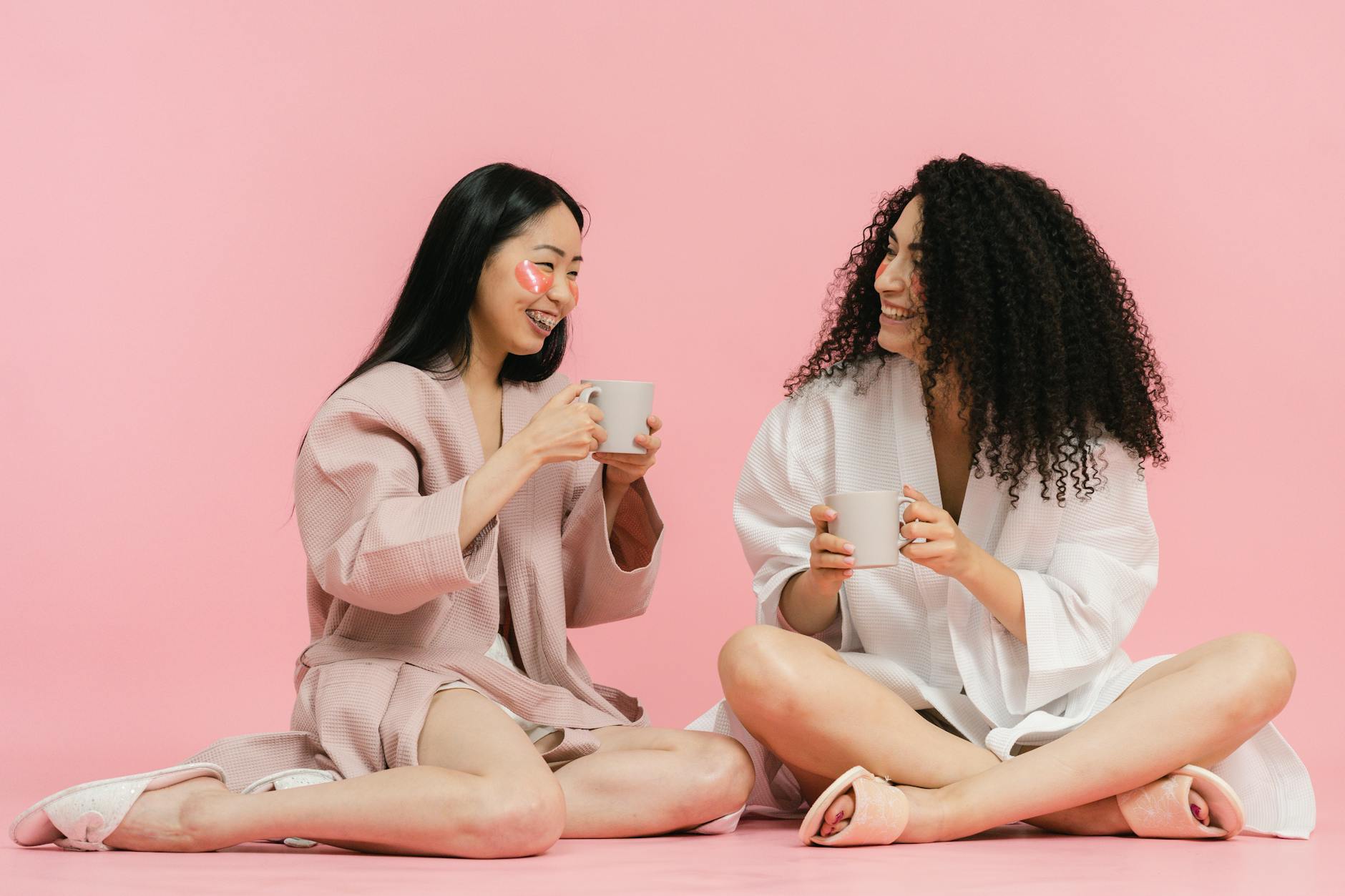 Two women in bathrobes relax with a warm drink, sitting on a pink background.