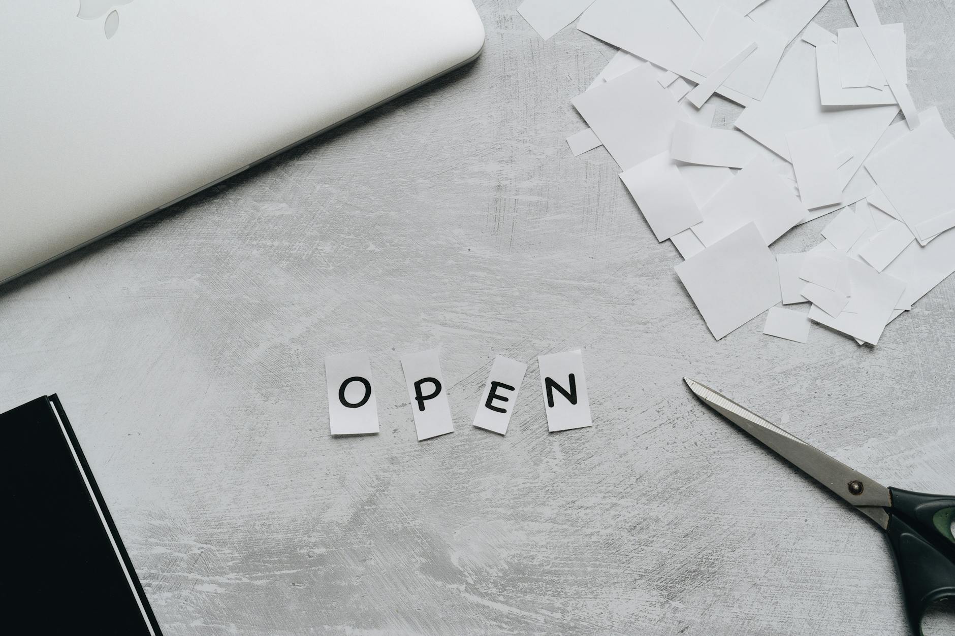 Flat lay of a creative workspace with paper letters spelling 'OPEN,' scissors, and a laptop.
