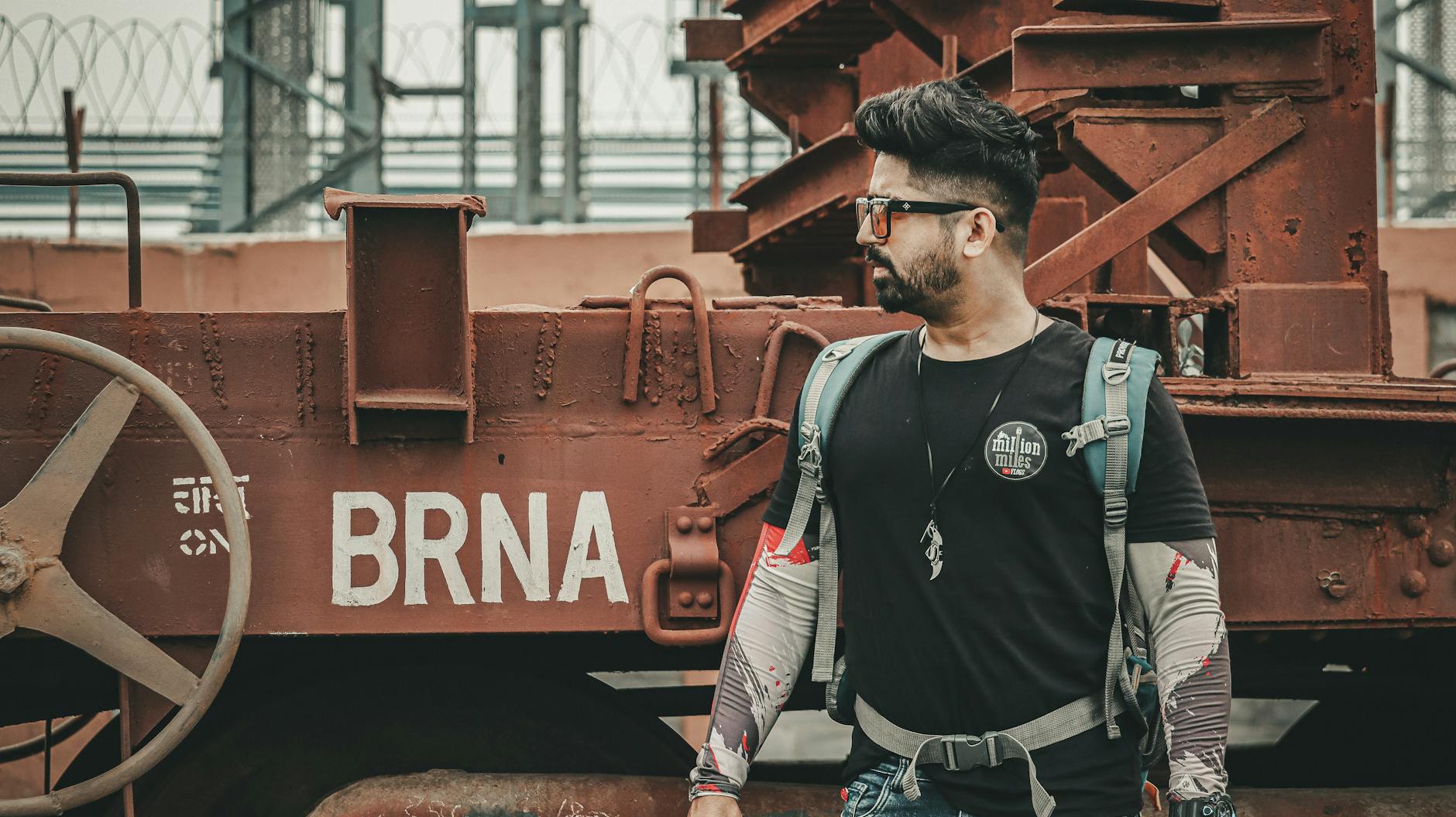 A fashionable man wearing sunglasses and a black shirt stands by a rusty train with 'BRNA' signage, portraying a modern traveler vibe.